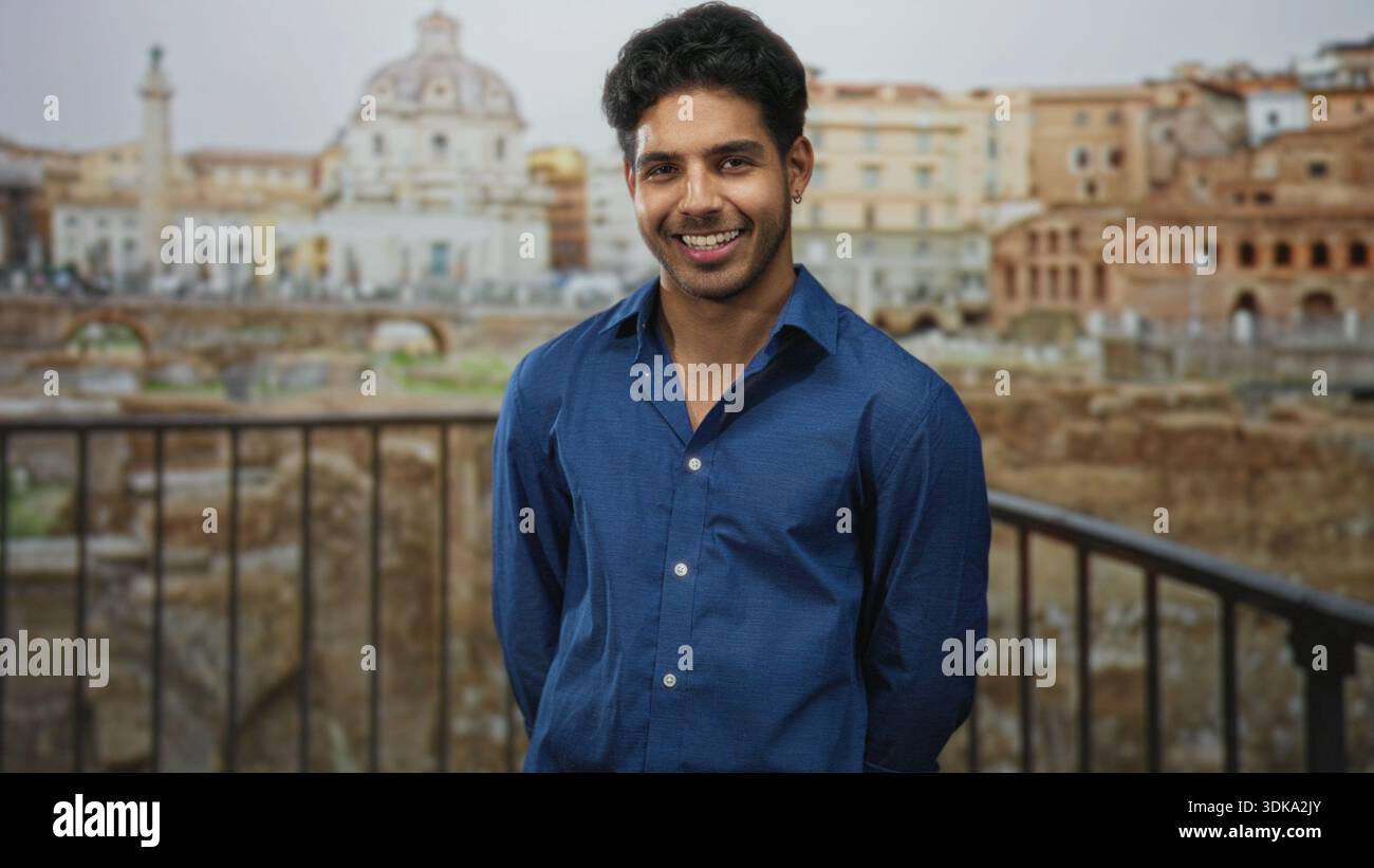 Uomo sorridente con le mani alle spalle appoggiato su ringhiere in metallo, indossa una camicia blu, si affaccia sulle antiche rovine romane e sull'edificio a cupola; gioioso viaggio mem Foto Stock
