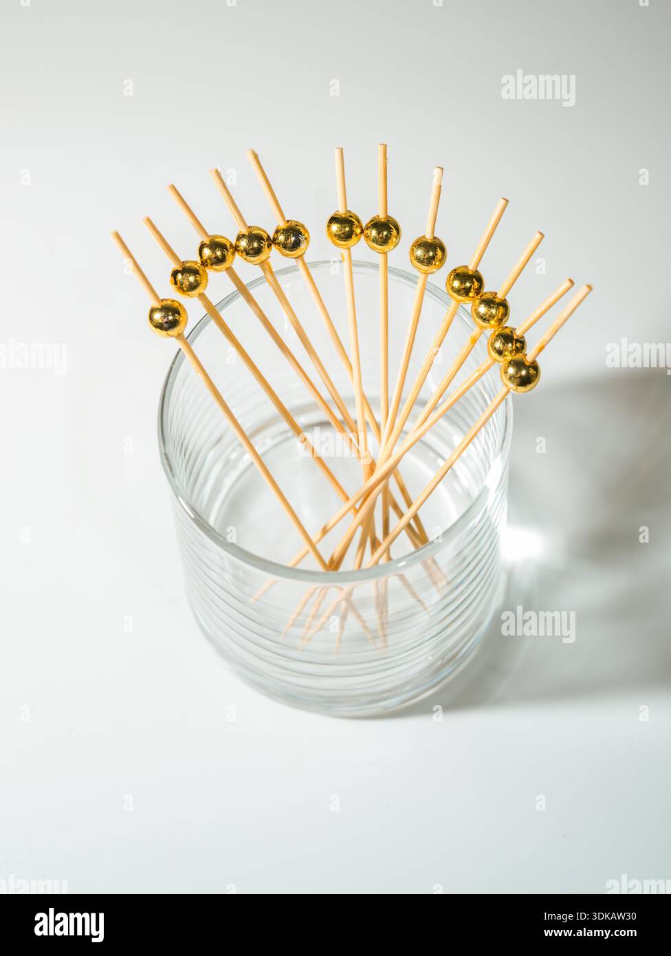 Una disposizione a ventaglio di spiedini da cocktail in bambù con eleganti perle dorate, in un bicchiere trasparente su una superficie bianca con har Foto Stock