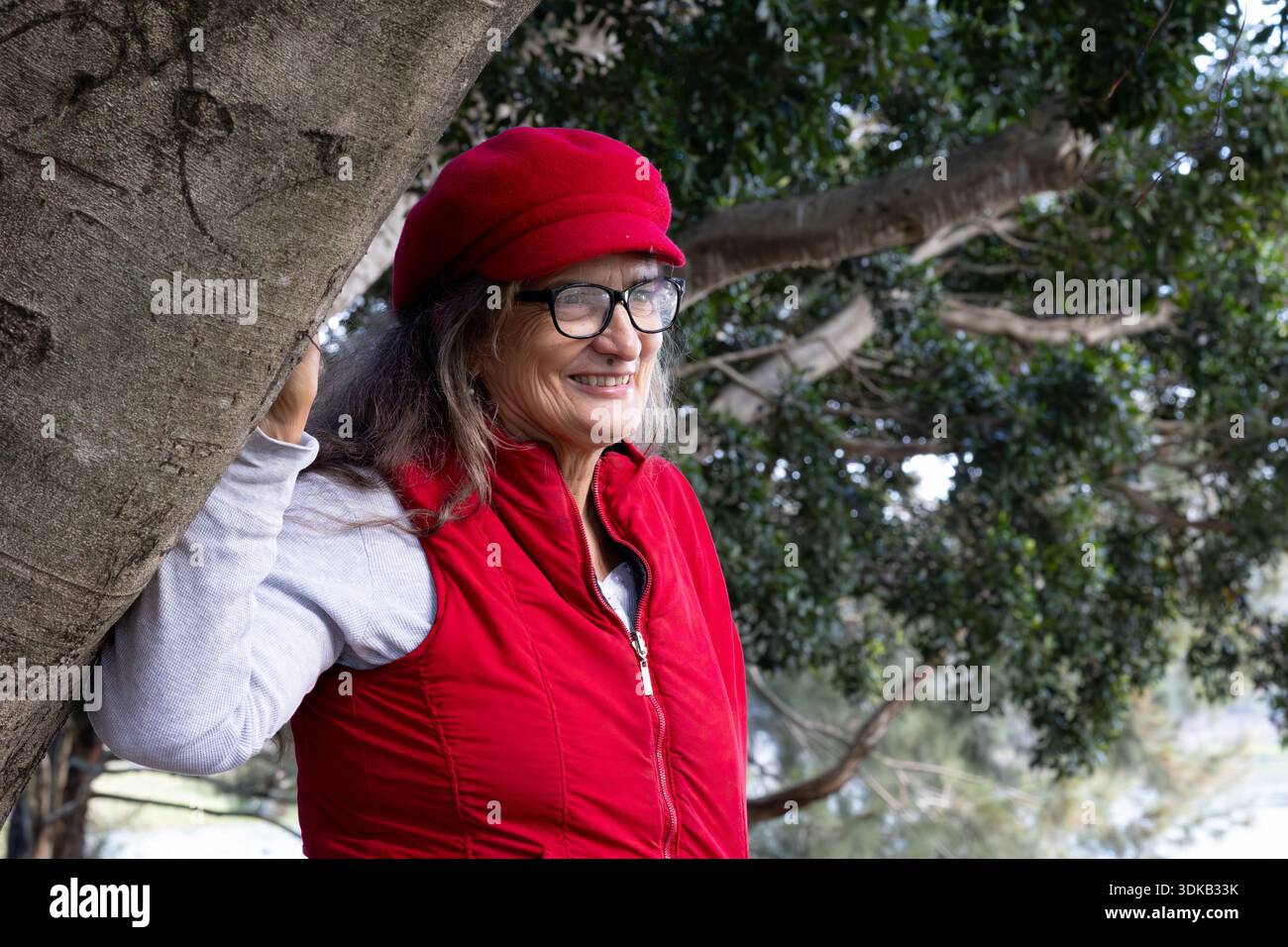 Una donna che indossa un gilet rosso e un berretto rosso sorride mentre riposa la mano su un ramo d'albero in un parco a Logan City, Queensland, Australia. Foto Stock