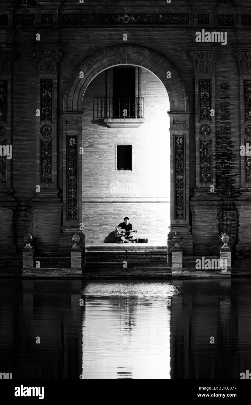 Fotografia in bianco e nero di una persona solitaria seduta sotto un monumentale arco di pietra riflesso nell'acqua ferma, creando un'immagine simmetrica e senza tempo Foto Stock