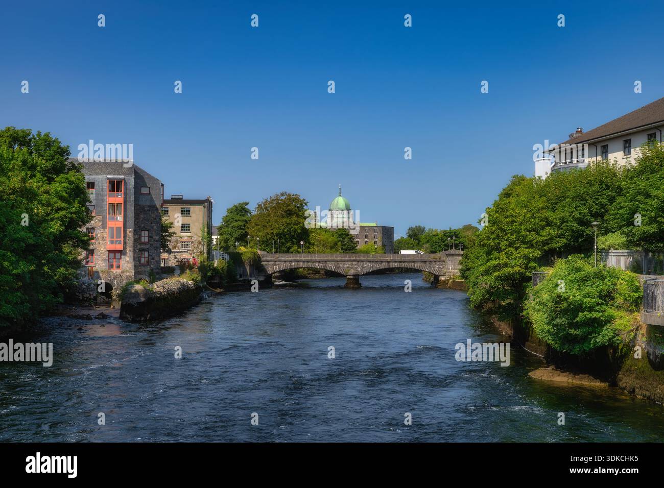 fiume illuminato dal sole con ponte in pietra, cupola storica scena lungo il fiume incorniciata da alberi frondosi ed edifici antichi, acque calme che riflettono il cielo limpido, fotografo di strada che cattura dettagli architettonici, Foto Stock