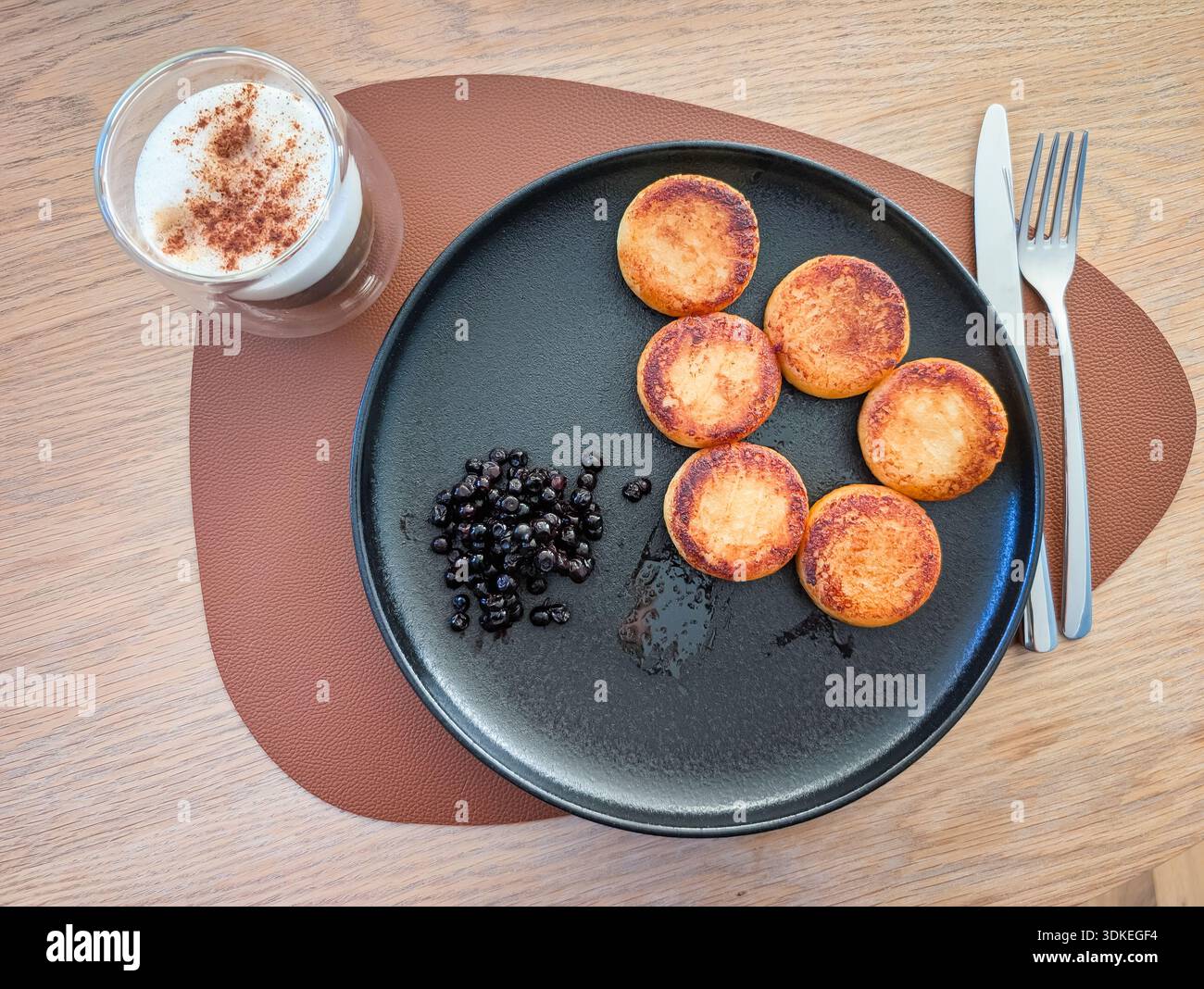 Deliziosa colazione a base di pancake fritti al formaggio servito con mirtilli su un piatto nero vicino al caffè con schiuma di cannella. Foto Stock