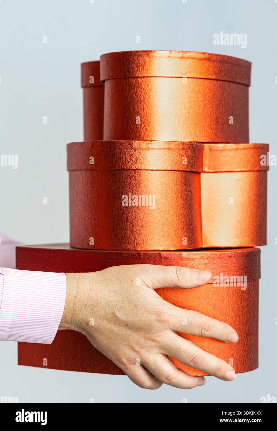 Primo piano di una mano con maniche rosa che bilanciano scatole regalo rotonde rosse-arancioni metallizzate sovrapposte, foto studio con spazio copia. Foto Stock