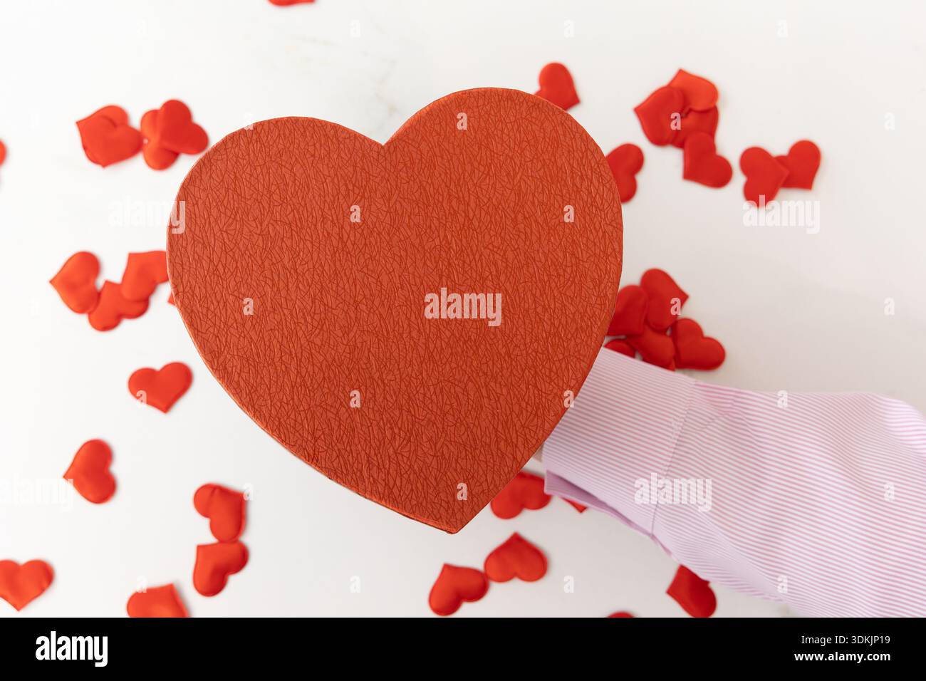 Colpo di mano verticale grandangolare a livello degli occhi con una camicia rosa che presenta una grande scatola a forma di cuore circondata da piccoli cuori rossi su una superficie luminosa Foto Stock