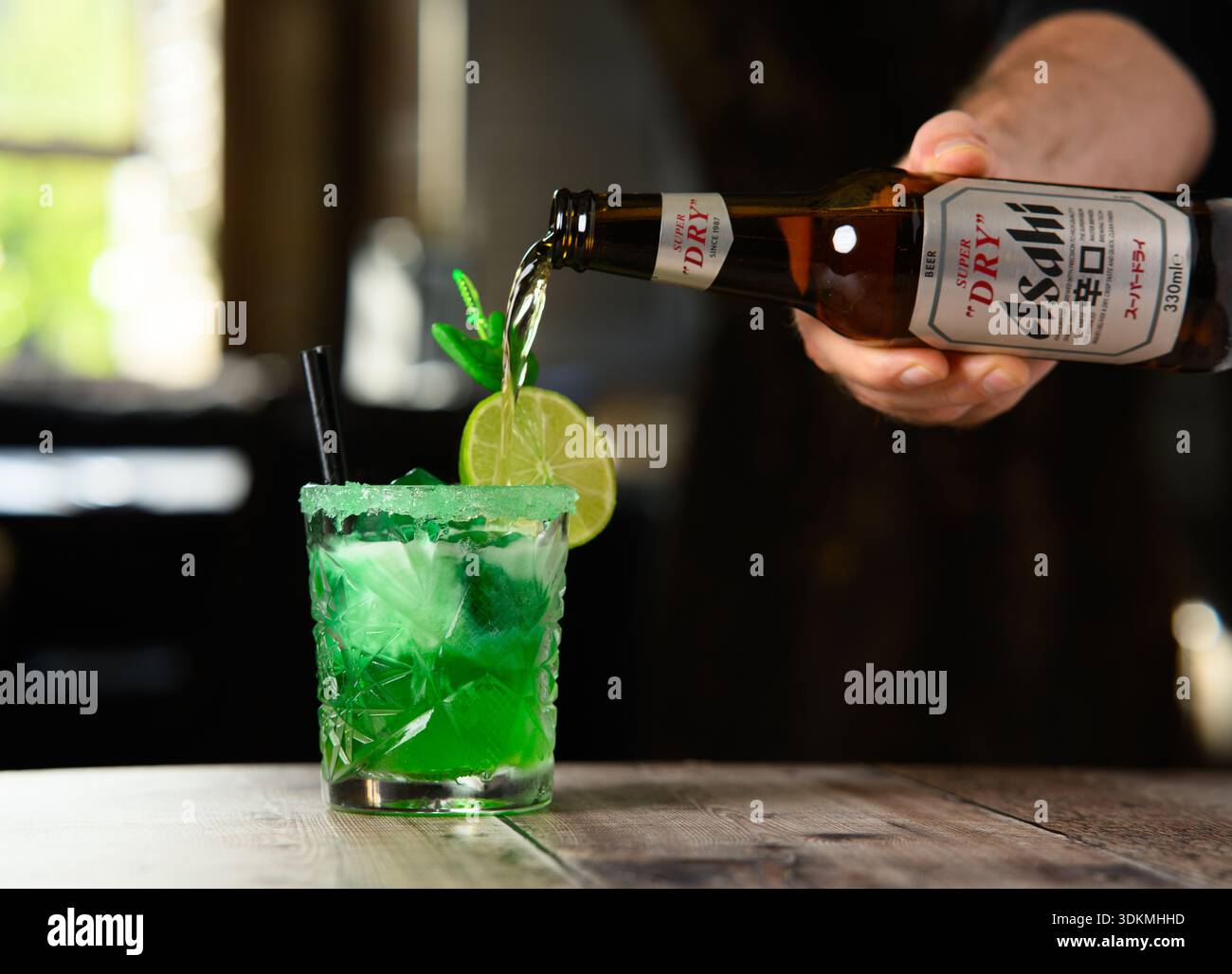 Un uomo sta versando una bevanda da una bottiglia in un bicchiere. La bevanda è verde e contiene un spicchio di lime Foto Stock