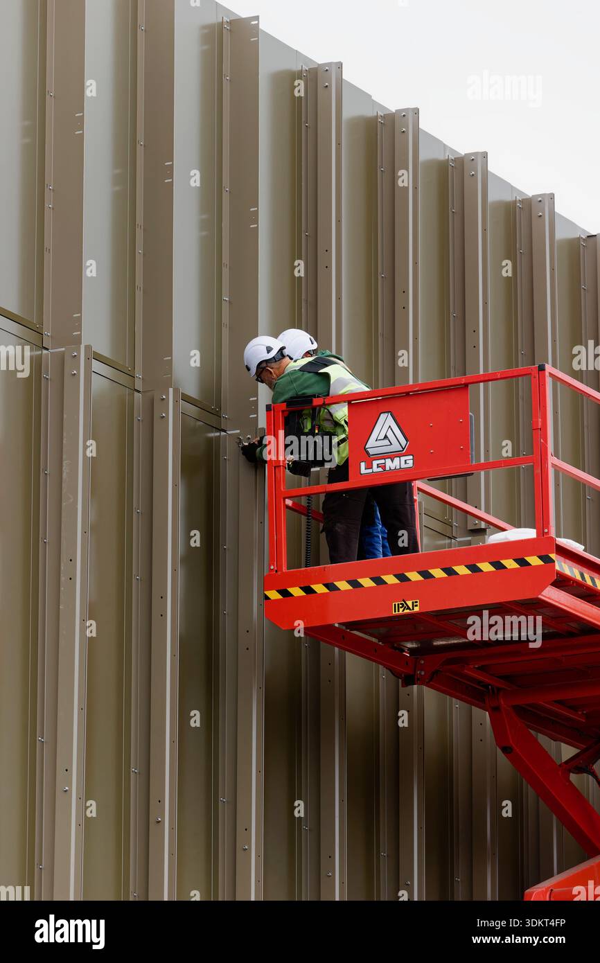 Due appaltatori con dpi sul sollevatore a forbice fissano le alette in alluminio ai pannelli di rivestimento verticali in alluminio sul mozzo civico radcliffe a Greater manchester regno unito Foto Stock