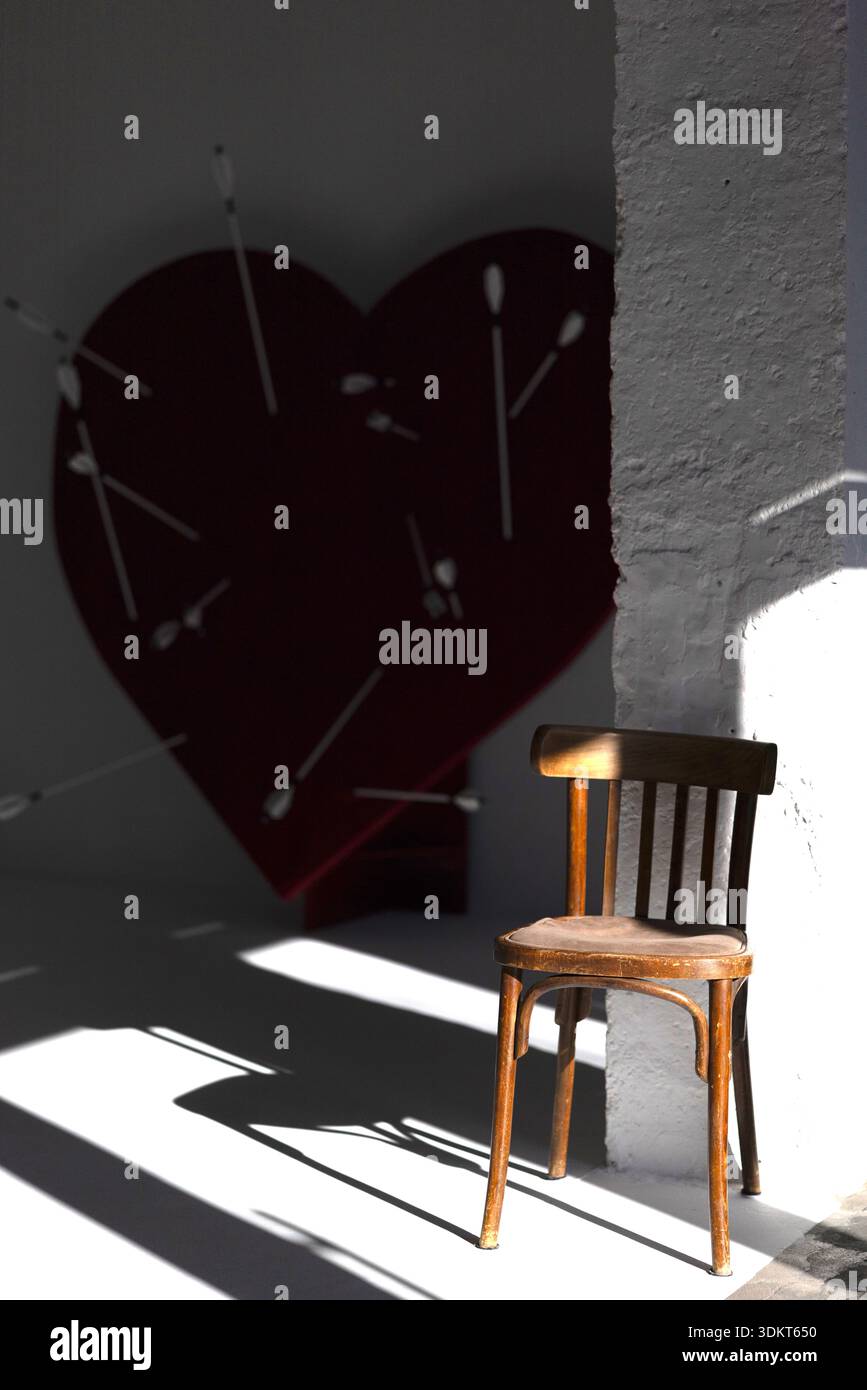 Ambiente interno minimalista con una sedia in legno con una suggestiva luce solare e una grande forma a cuore con frecce visibili in ombra sulla parete. Concetto di amore, Foto Stock