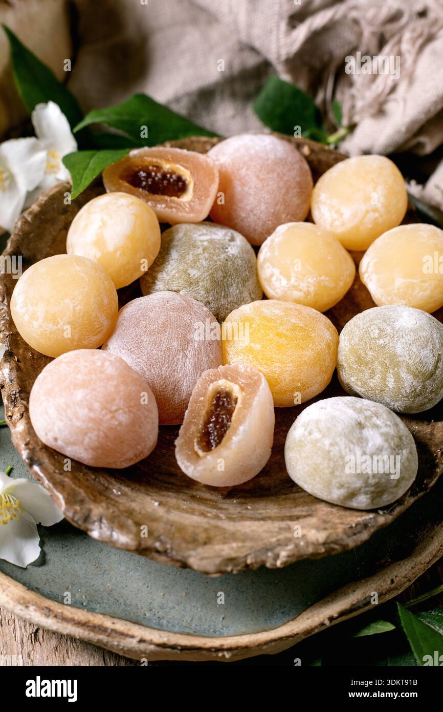 Colorate torte di riso mochi tradizionali giapponesi con ripieno dolce su un piatto rustico di legno decorato con fiori bianchi e foglie verdi Foto Stock