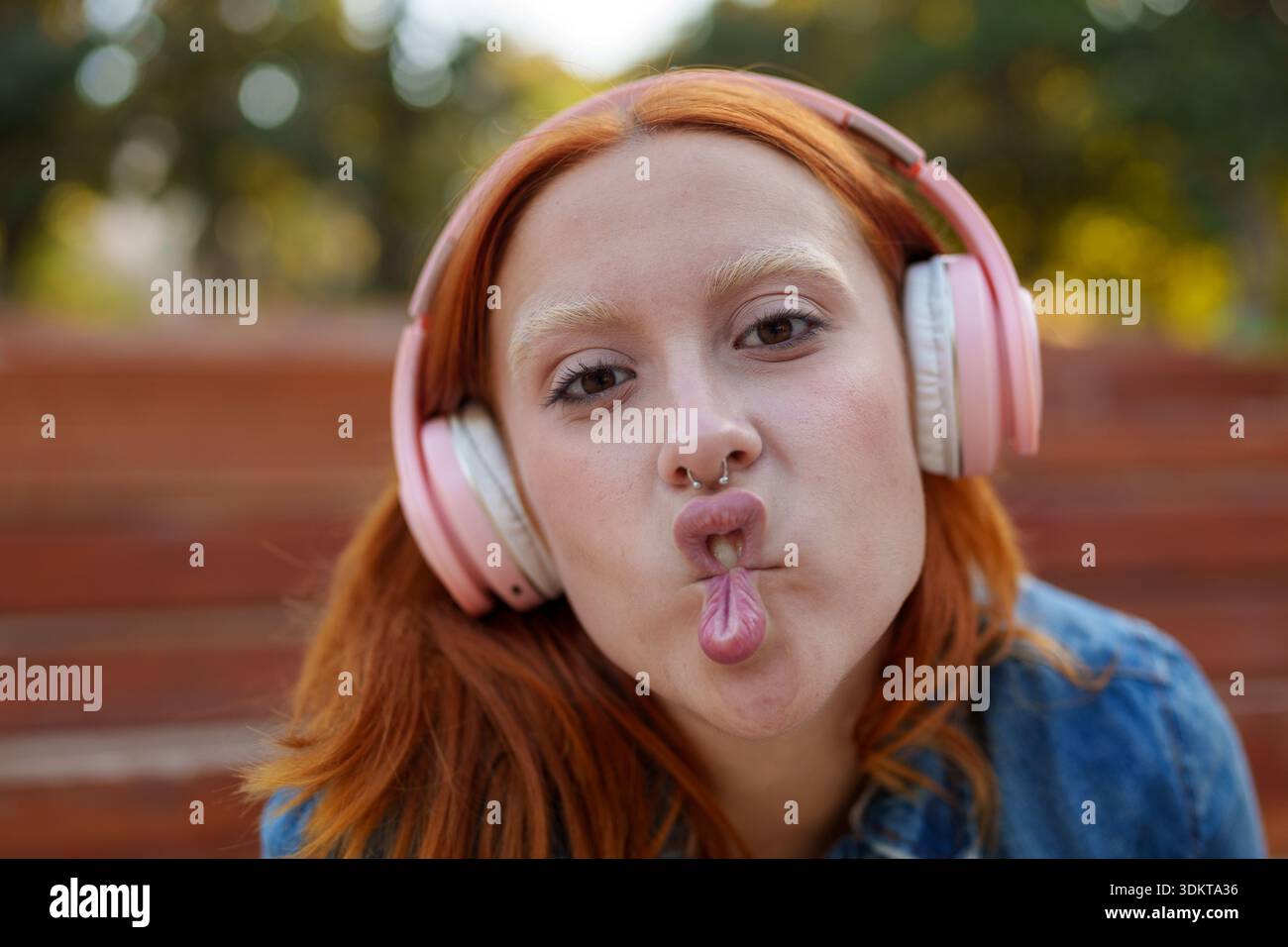 Giovane donna giocosa con i capelli rossi vivaci e una piercing, che posa e assorbe la musica in un ambiente all'aperto Foto Stock