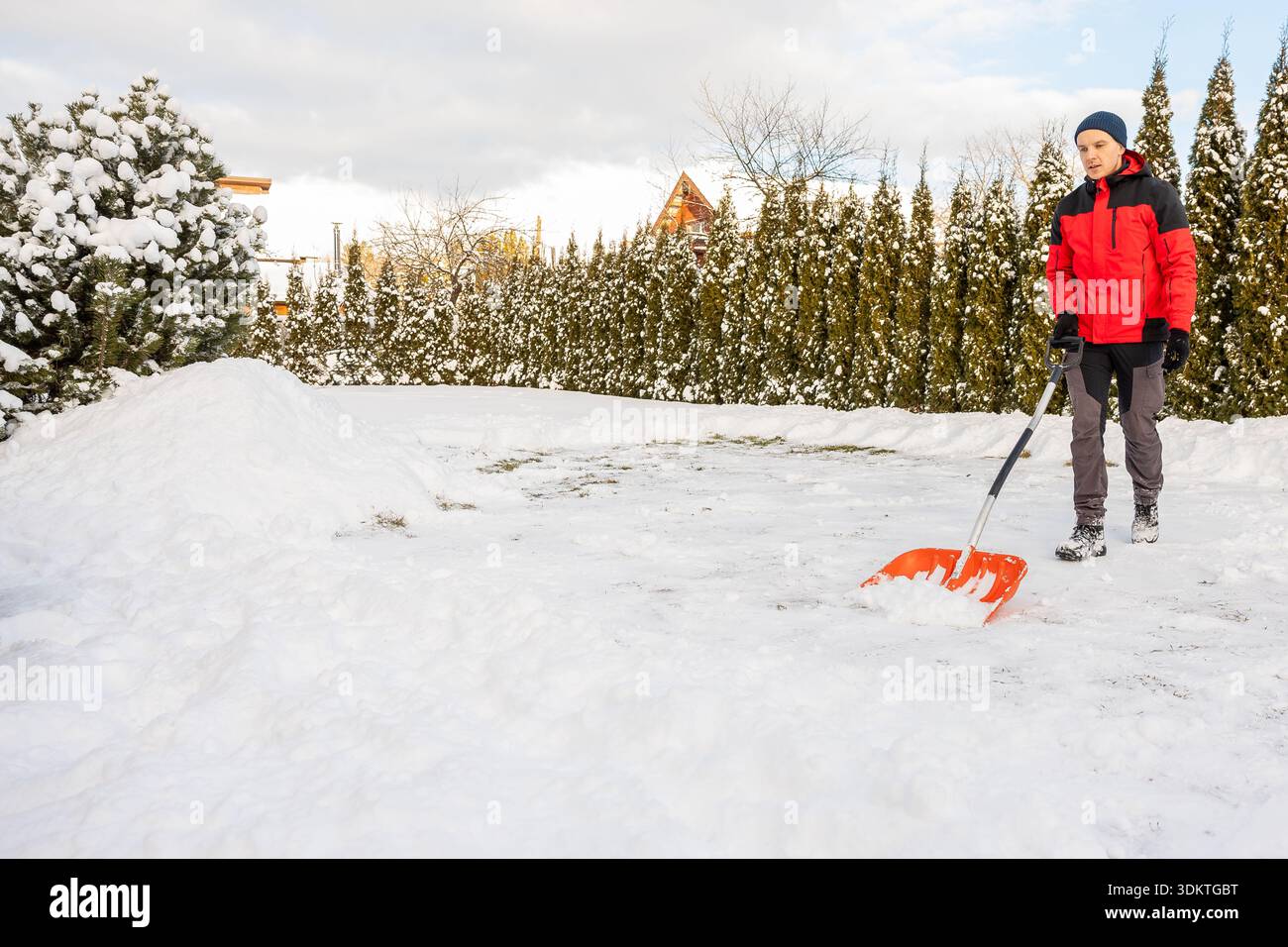 Indossato in giacca rossa e pantaloni grigi, pulisce la neve su un sentiero circondato da alti sempreverdi, cespugli ricoperti di neve e una struttura a tetto triangolare. Foto Stock