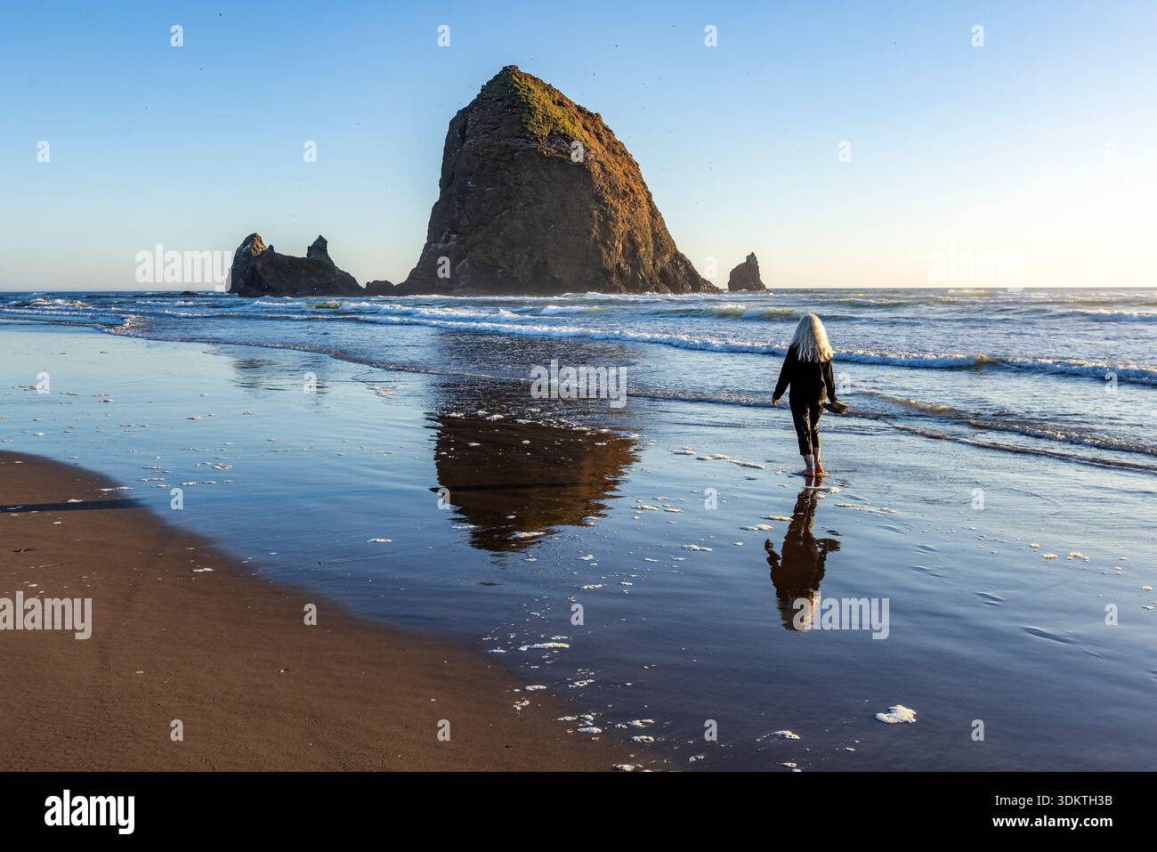 USA, Oregon, Cannon Beach, Vista posteriore di una donna che cammina sulla spiaggia vicino alla roccia del pagliaio Foto Stock