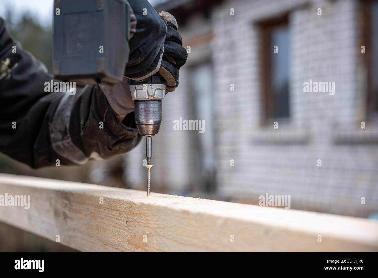 Una persona utilizza un trapano elettrico per inserire una vite in una tavola di legno, con le mani dei guanti visibili. Sullo sfondo appare un edificio in mattoni con finestre. Foto Stock