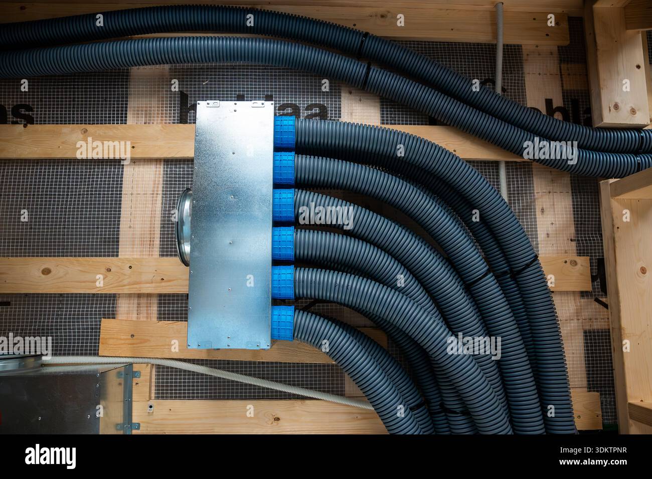 Vista dettagliata dei condotti corrugati neri e della scatola di distribuzione in metallo con connettori blu in una struttura in legno, con barrie in vapore a griglia Foto Stock