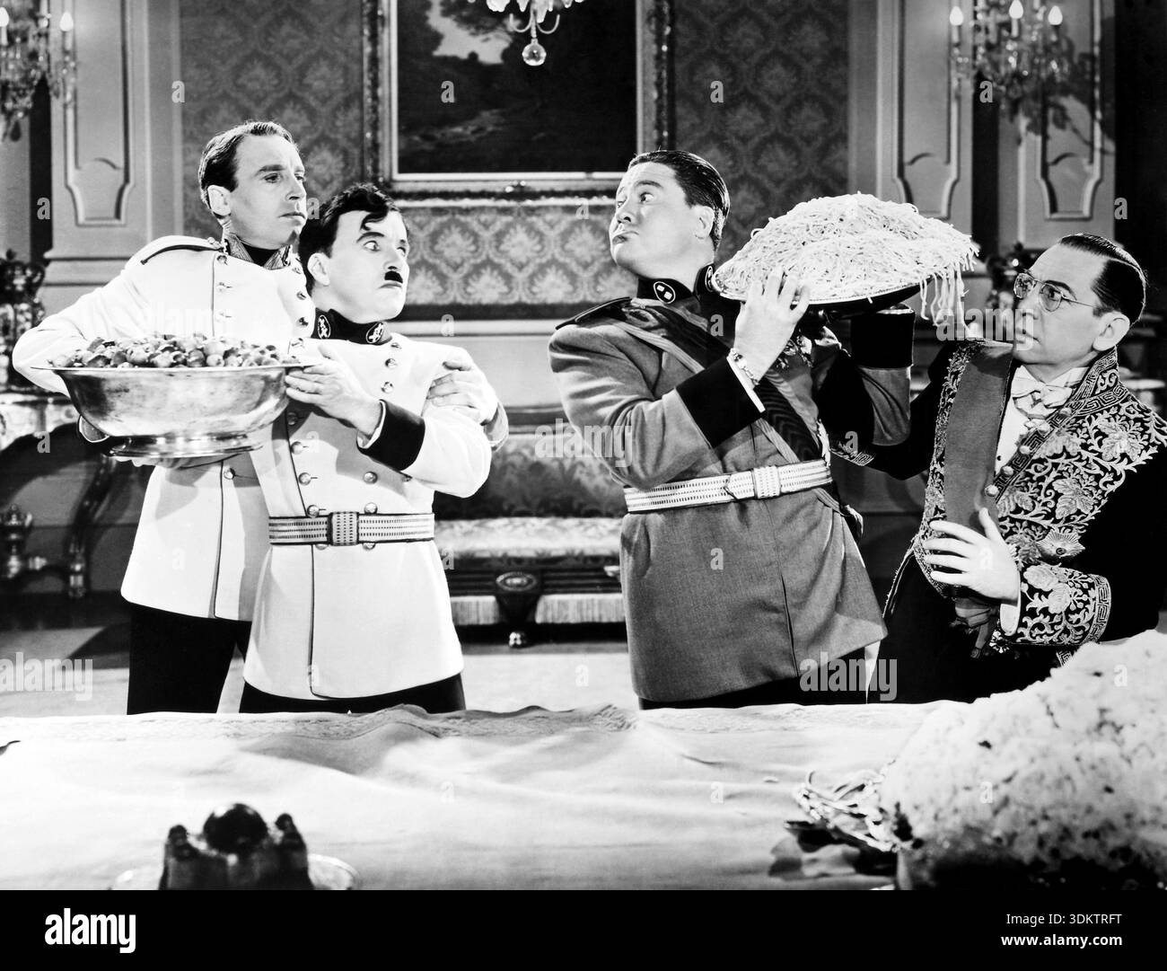 Questo iconico bianco e nero ancora tratto dal film del 1940 "il grande dittatore" cattura un momento comico con Charlie Chaplin, che interpreta l'adenoide Hynkel, insieme a personaggi come Jack Oakie nel ruolo di Benzino Napaloni e Henry Daniell nel ruolo di Garbitsch. La scena li ritrae in modo umoristico interagendo con grandi ciotole di cibo, una piena di frutta e l'altra con un tumulo di spaghetti a forma di cappello. Riflette l'interpretazione satirica del film sui regimi totalitari della sua epoca. Foto Stock