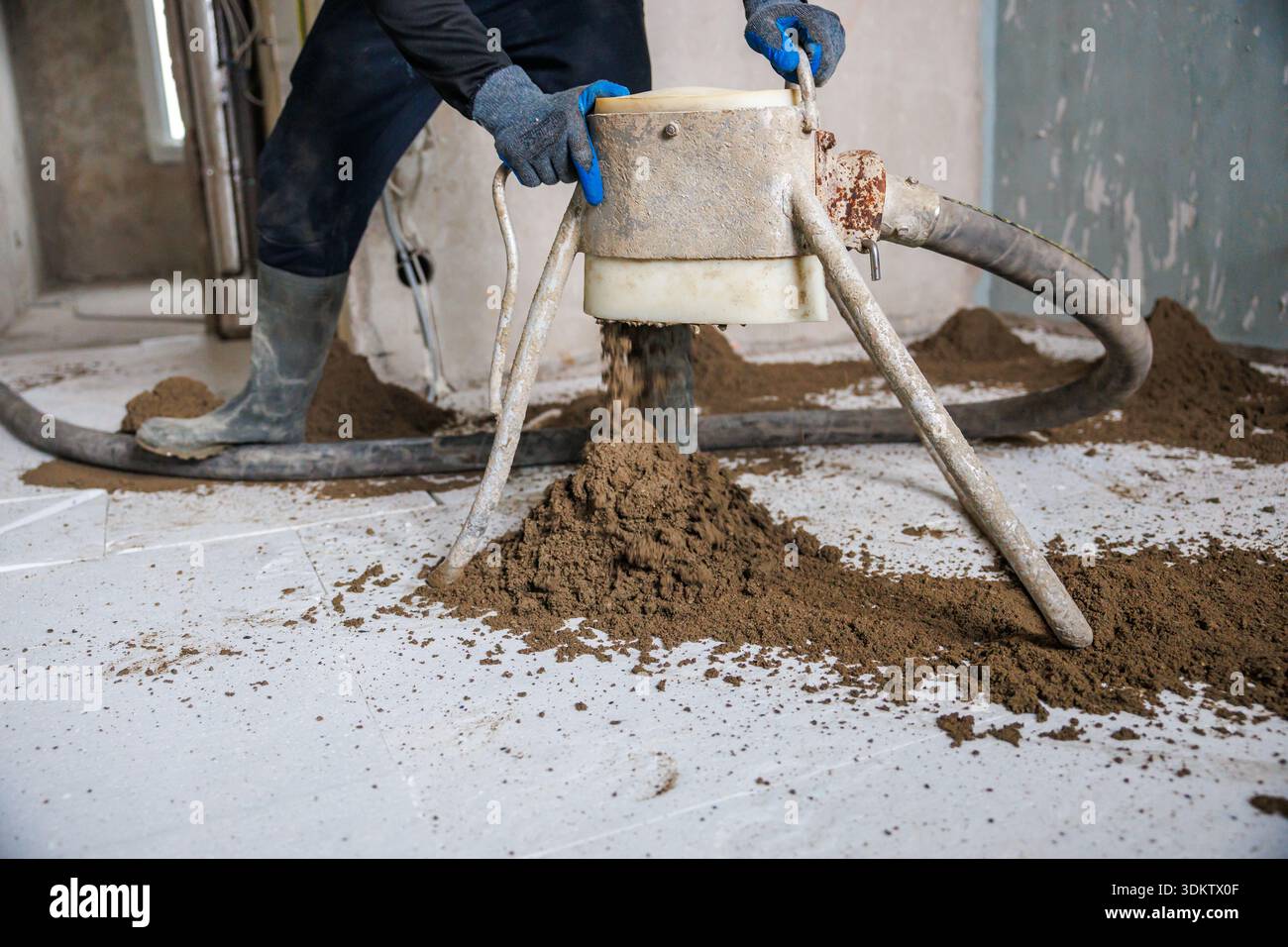 Un lavoratore con guanti e stivali utilizza uno strumento con gambe di treppiede e un tubo flessibile, formando pile di sabbia o terreno su un pavimento piastrellato in un sito interno. Foto Stock