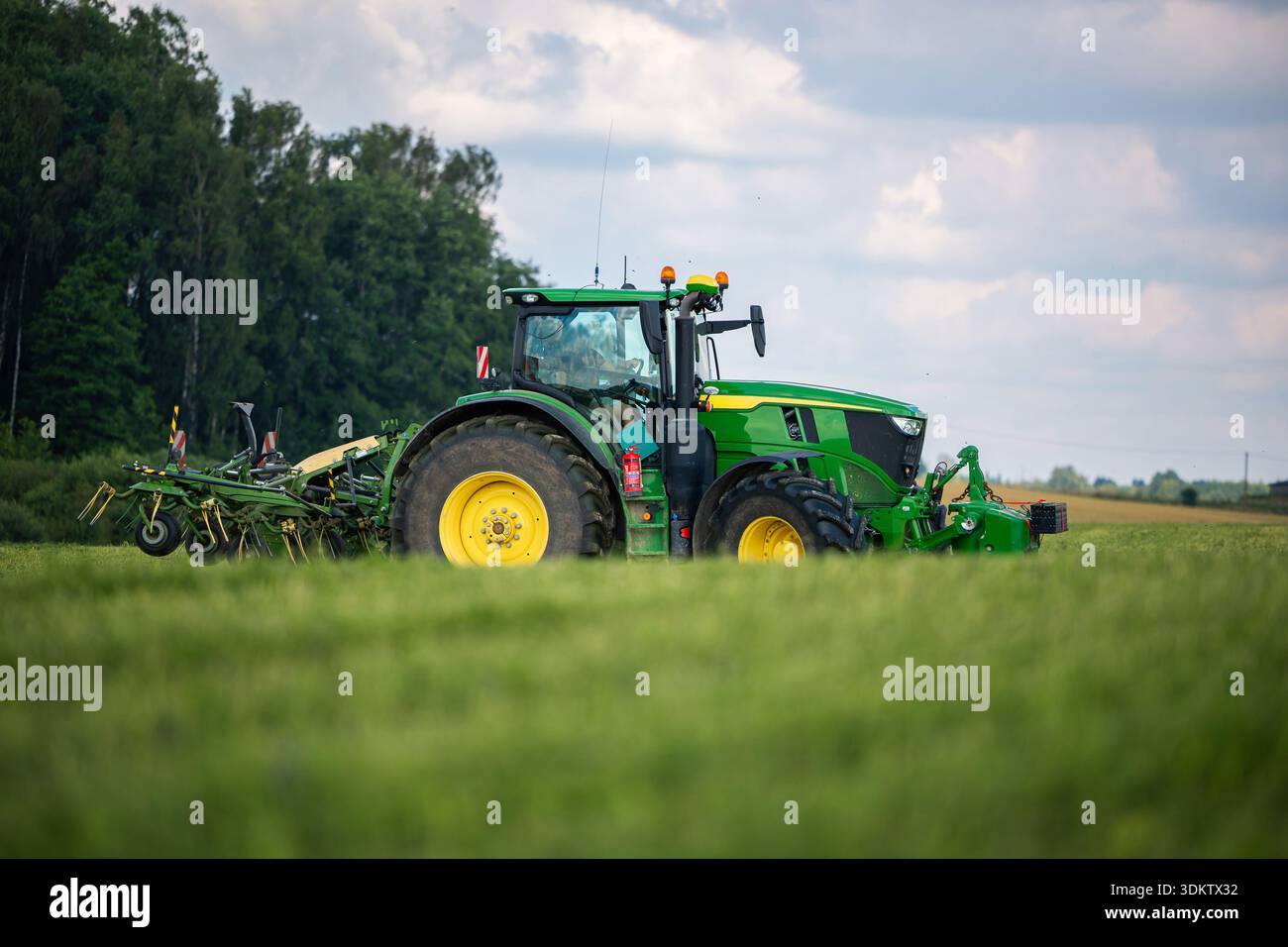 Il trattore verde e giallo con pneumatici grandi tira un attrezzo multicolore attraverso l'erba alta in terreni agricoli aperti. La cabina mostra specchietti, luci e a. Foto Stock