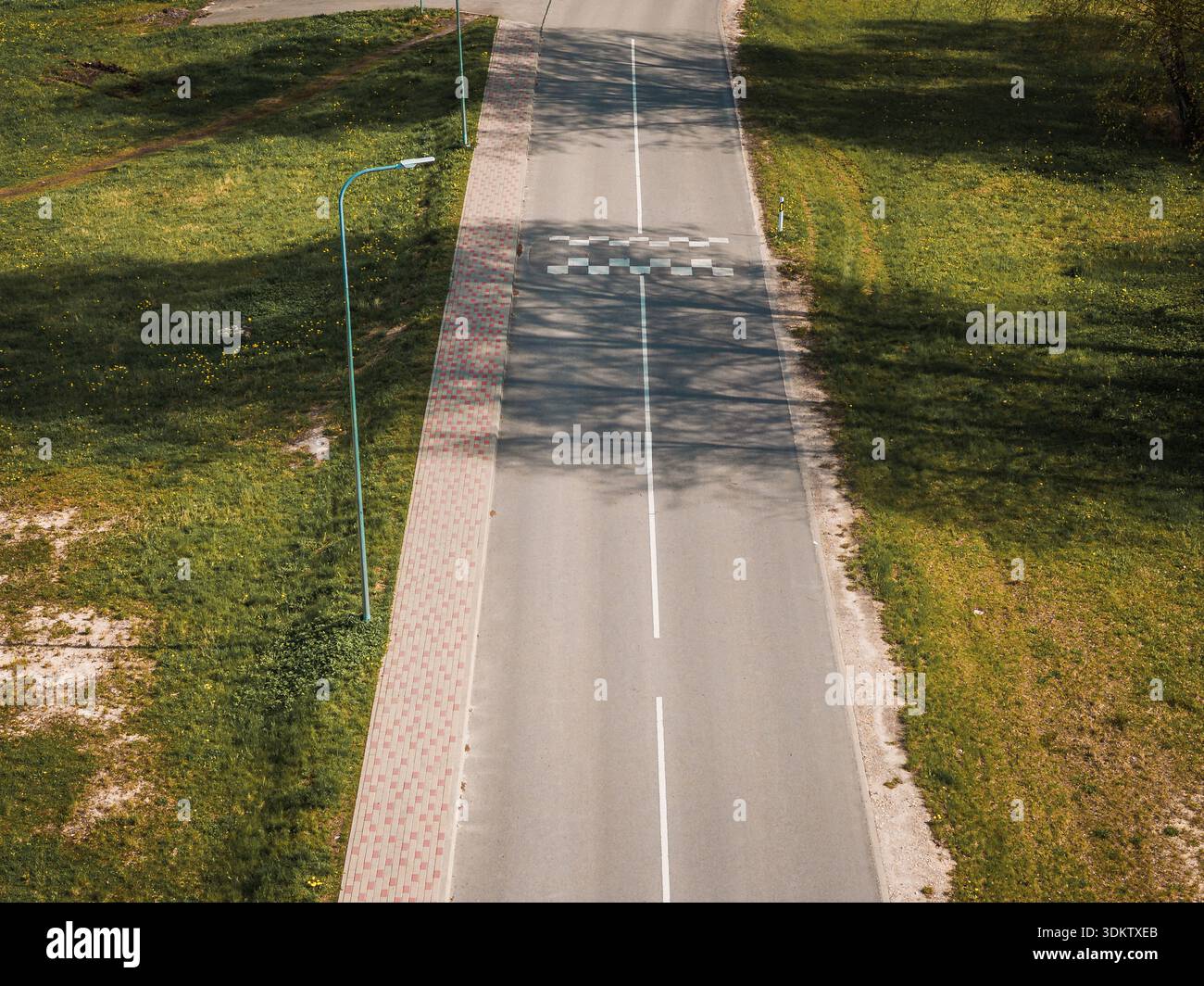 Una strada asfaltata dritta con un passaggio pedonale segnalato, erba su entrambi i lati, un lampione sulla sinistra e un marciapiede pavimentato in mattoni parallelo al roa Foto Stock