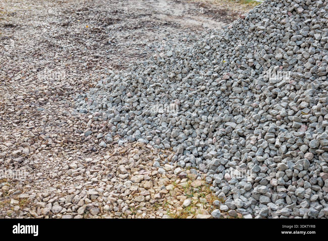 Un grande mucchio di piccole pietre grigie poggia su terra irregolare e ghiaia, circondato da rocce sparse e macchie d'erba, suggerendo un cantiere edile. Foto Stock