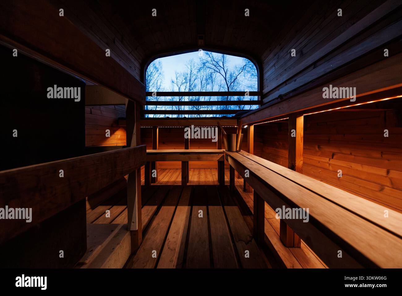 Interno sauna a botte con panche in legno a gradini, secchio in mestolo metallico e illuminazione a strisce. Le silhouette in betulla senza foglie appaiono al crepuscolo, per un contrasto caldo e fresco Foto Stock