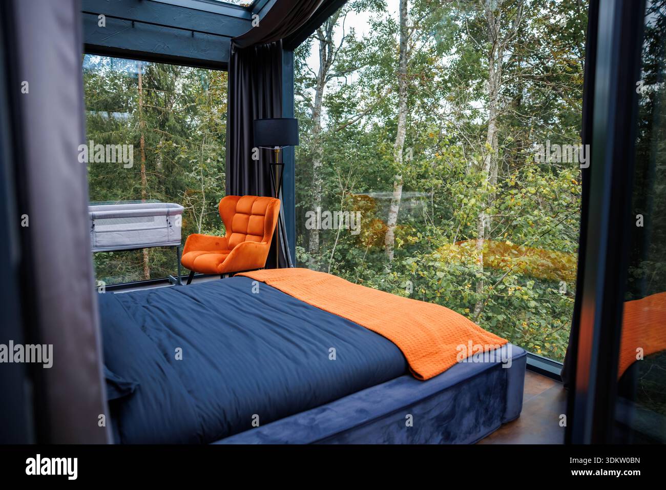 Una camera moderna con pareti e soffitto in vetro, con letto blu navy, accenti arancioni e vista su alti alberi in un'area boschiva. Foto Stock