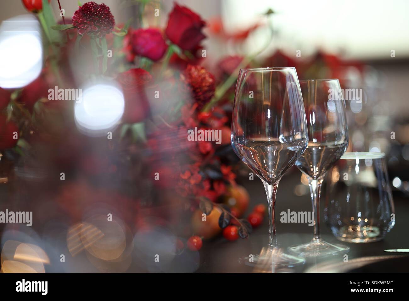 Bicchieri da vino in cristallo vuoti che riflettono la luce sul tavolo scuro, caratterizzati da un bellissimo centro floreale rosso che crea un'atmosfera da festa, raffinata Foto Stock