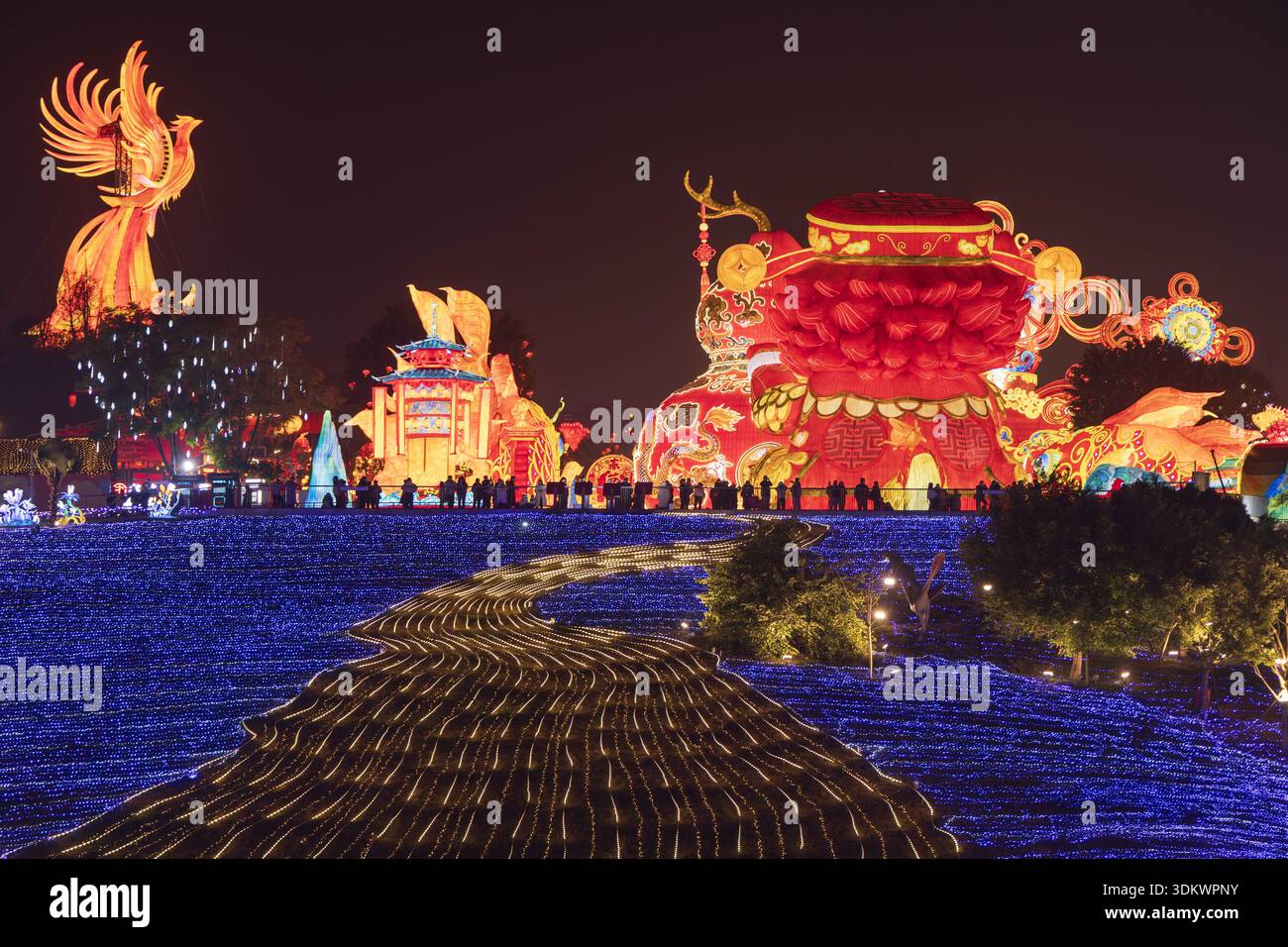 Vista di un fiume luminoso di luce blu che si snoda attraverso un paesaggio vivace punteggiato da luminescenti fenici, lanterne ornate e sagome di persone Foto Stock
