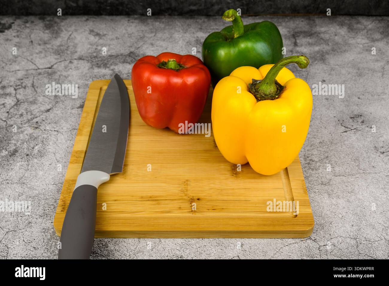 Tagliere in legno con coltello da cuoco e peperoni freschi (rosso, verde, giallo) su sfondo grigio. Preparazione minima di cottura, cibo sano, primavera. Foto Stock