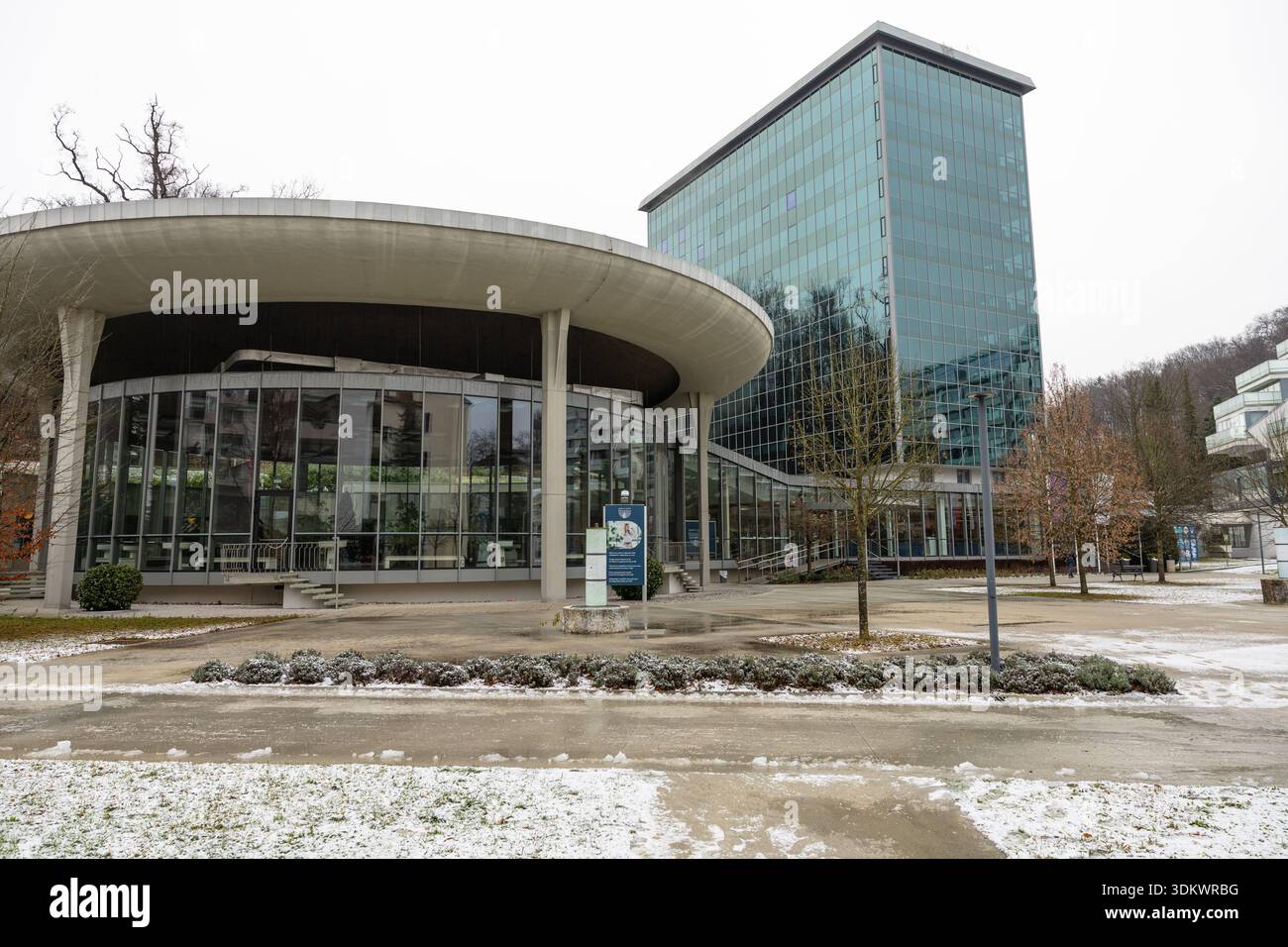 ROGASKA SLATINA, SLOVENIA, 01.03.2026: Un moderno complesso di uffici in vetro caratterizzato da un baldacchino curvo e da un'alta torre su un campus innevato. Ideale per archi Foto Stock