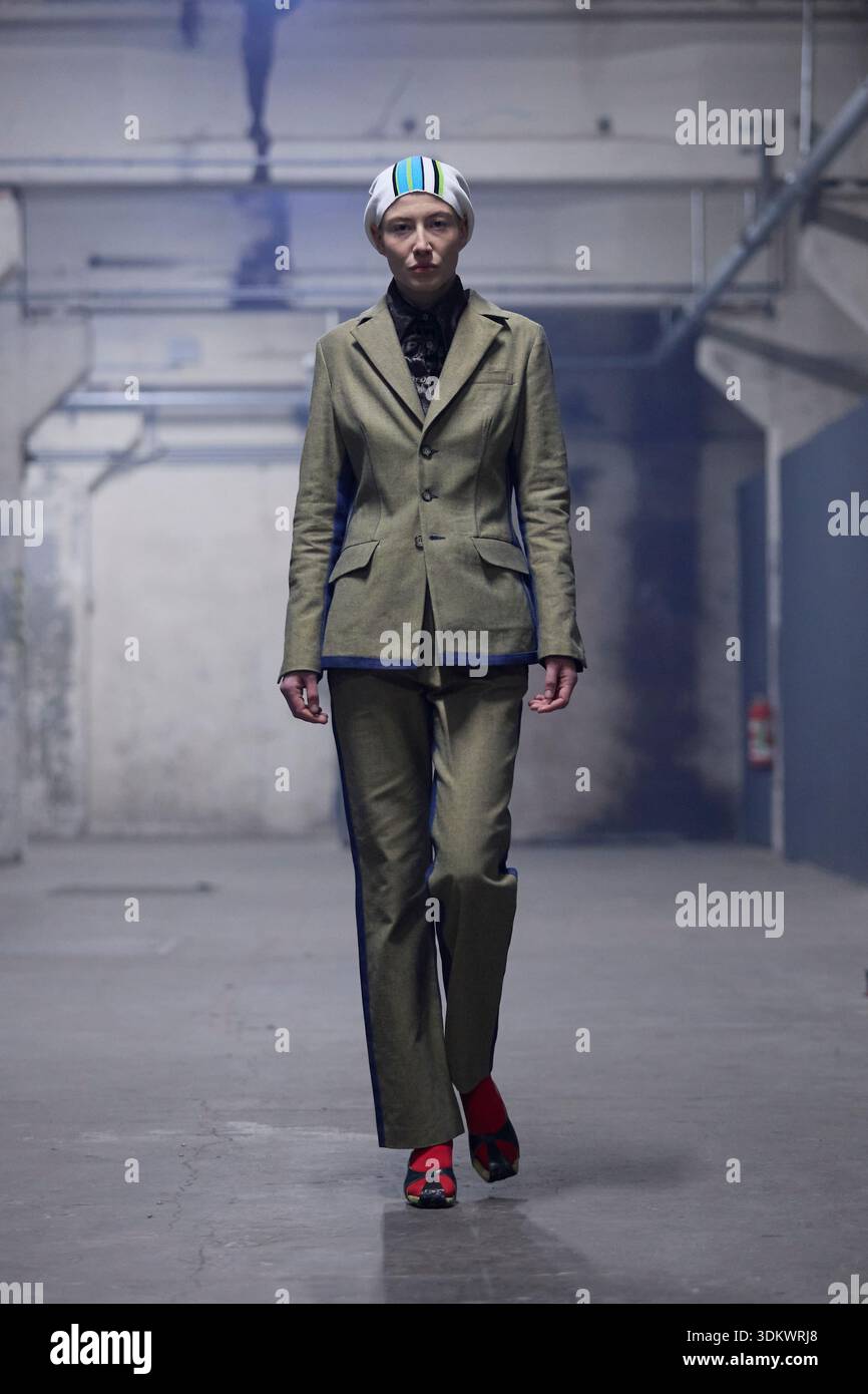 Berlino, Germania, 2 febbraio 2026. Una modella percorre la passerella della collezione autunno/inverno 2026 di Kenneth Ize durante la Berlin Fashion Week a Kraftwerk. Il look sartoriale è caratterizzato da un blazer monopetto verde oliva sartoriale con finitura blu lungo l'orlo inferiore, abbinato a pantaloni verdi coordinati. L'outfit è completato da una camicia a motivi scuri, un copricapo a righe azzurro e bianco e scarpe rosso brillante, che mostrano l'attenzione del designer verso l'elegante sartoria e dettagli artigianali sotto l'illuminazione industriale. Crediti: Felix Wolf/Alamy Live News Foto Stock