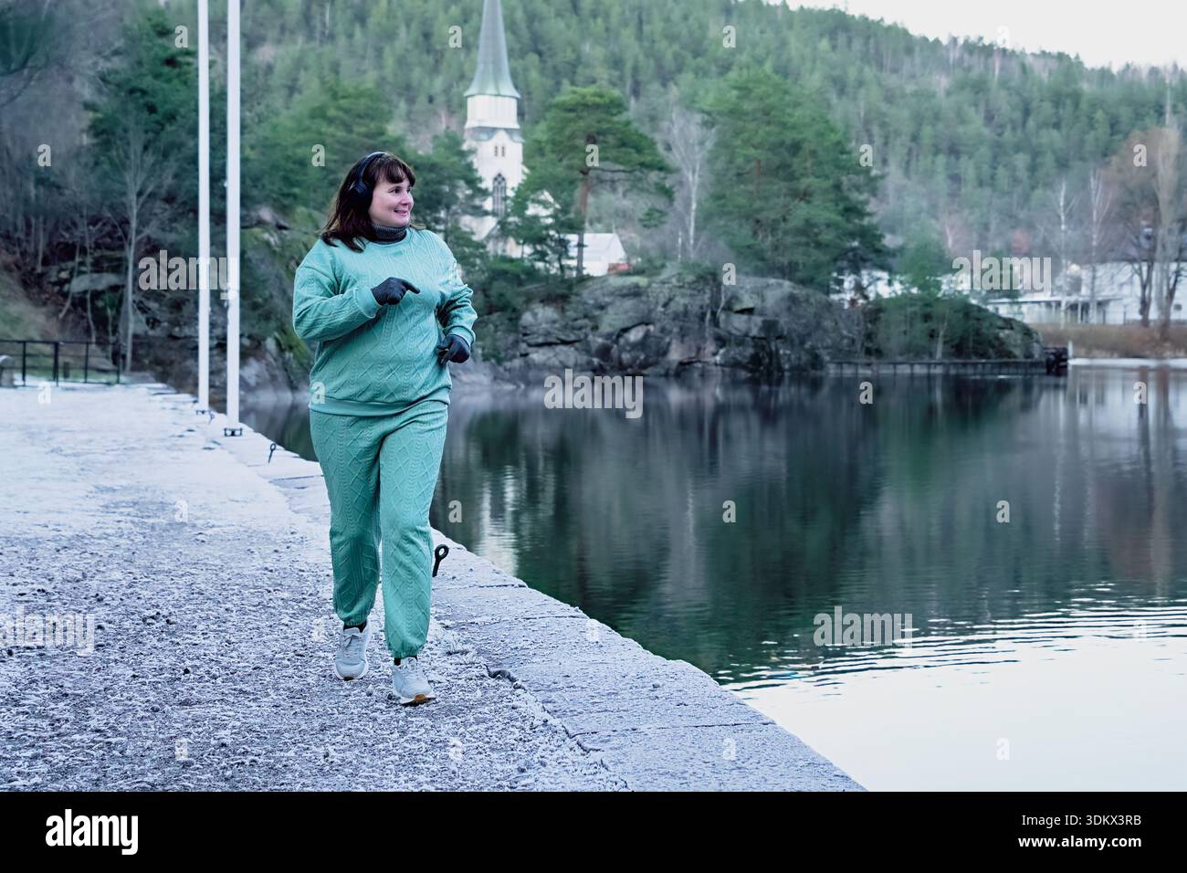 Una jogger più grande in costume color tè corre lungo un tranquillo sentiero sul lago, con alberi e un lontano campanile della chiesa, che incarna salute e all'aperto acti Foto Stock