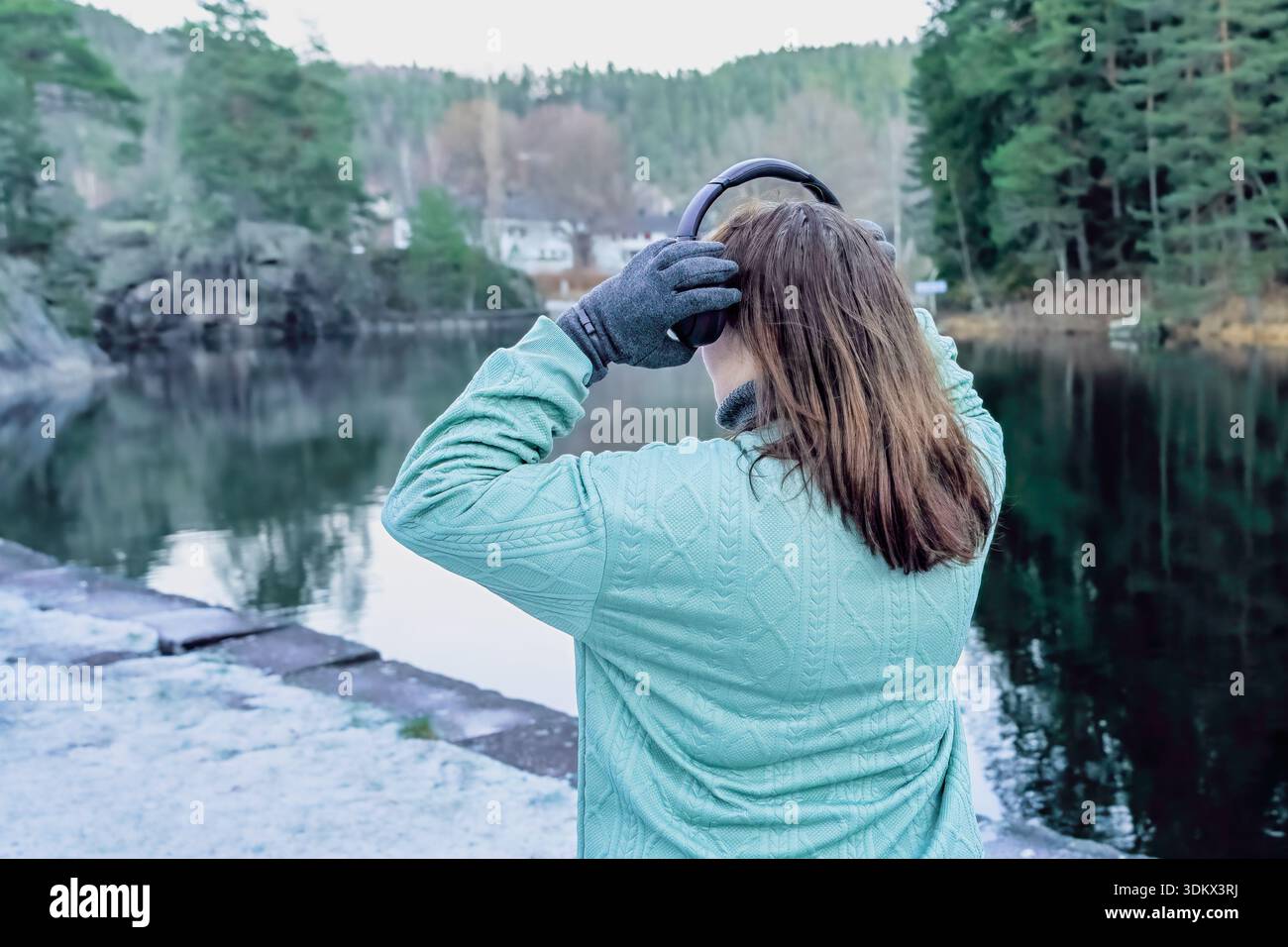 La figura femminile con le cuffie regola i capelli mentre si trova in riva a un lago tranquillo, circondato da una vegetazione lussureggiante, creando un'atmosfera calma e riflessiva Foto Stock