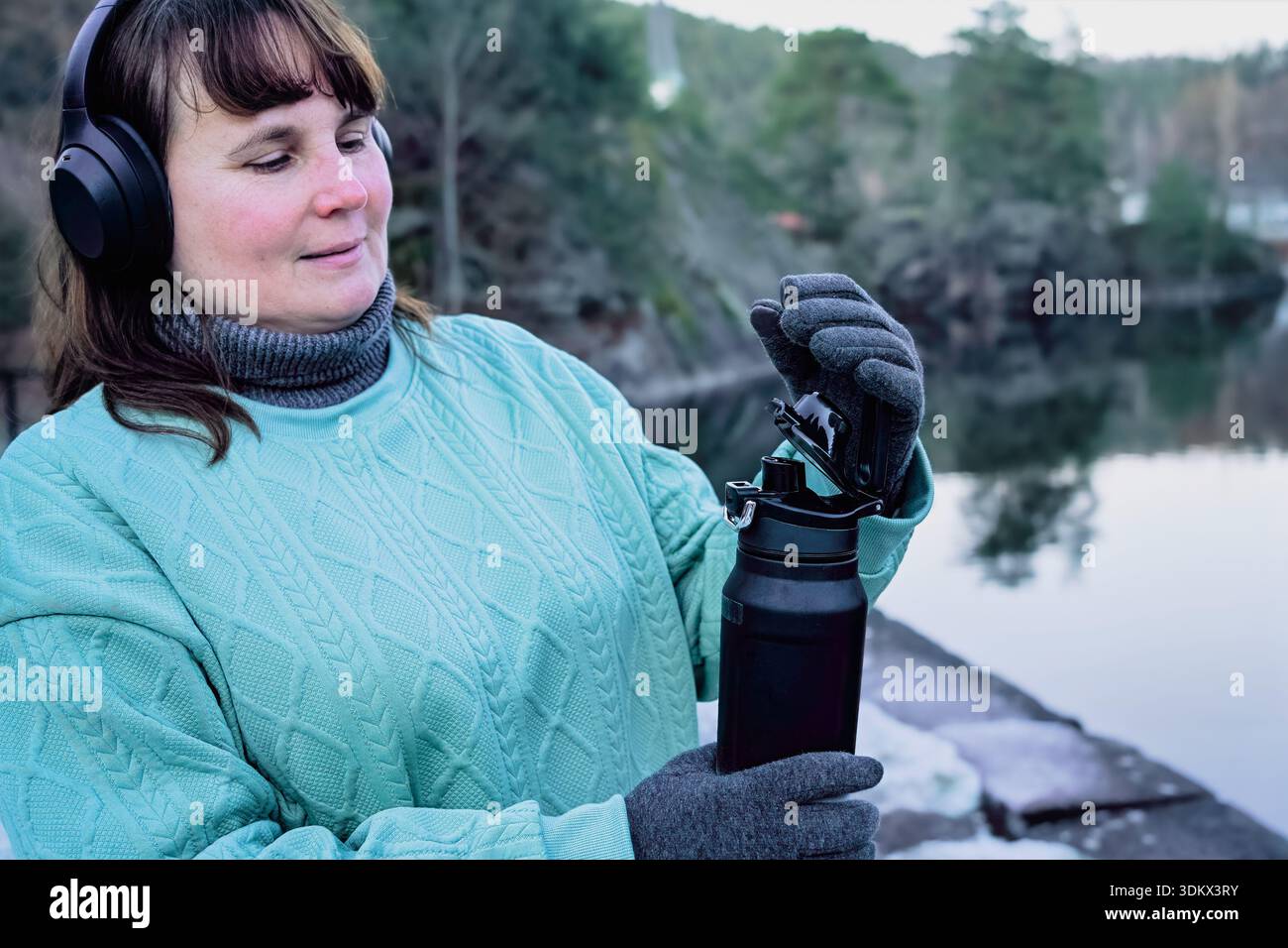 Donna che ama la natura, aprendo una bottiglia d'acqua mentre indossa le cuffie e un maglione caldo, in piedi accanto a un tranquillo lago con alberi sullo sfondo, em Foto Stock