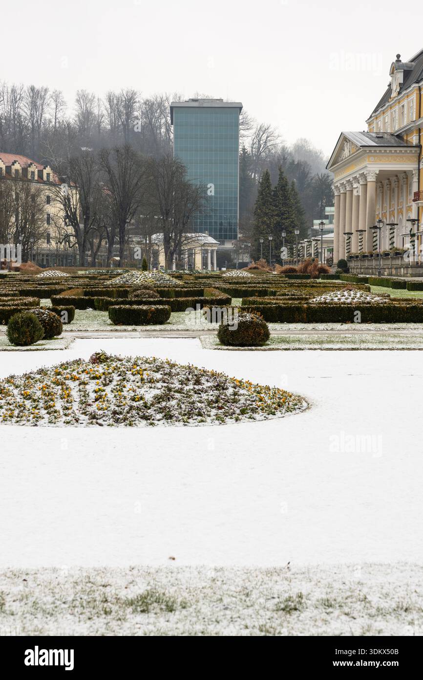 Un tranquillo ambiente invernale caratterizzato da un giardino formale con siepi rifinite, aiuole spolverate di neve, un grandioso edificio neoclassico giallo e un elegante vetro Foto Stock