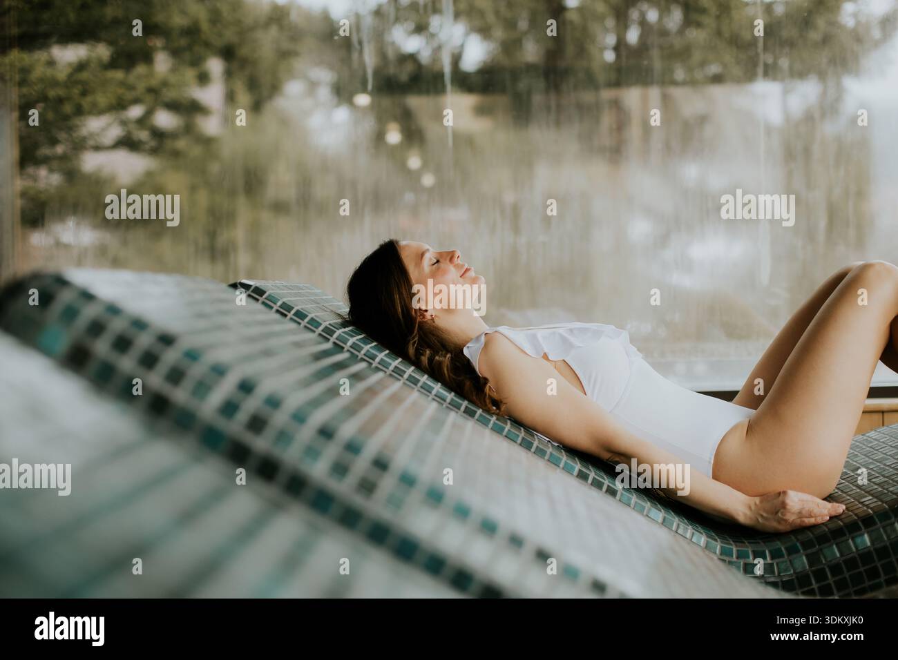 Una donna giace su una poltrona piastrellata accanto ad un elemento acquatico. Si gode un momento di riposo mentre la luce del sole filtra attraverso lo spazio. Il settin Foto Stock