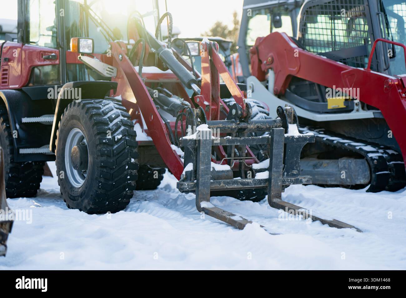 Forche per movimentazione materiali su pala rossa nei cantieri innevati, con pala cingolata compatta nelle vicinanze, che cattura la gamma di attrezzature per l'edilizia invernale per il groun congelato Foto Stock