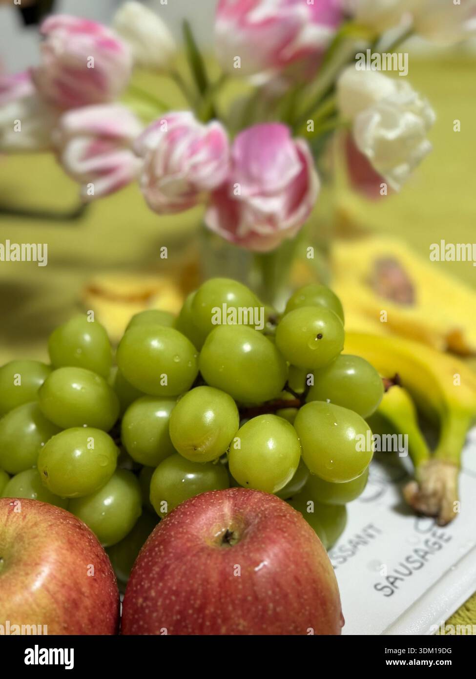Frutta fresca natura morta con uva verde ricoperta di rugiada, mele rosse, banane e delicato bouquet di tulipani rosa-bianco su sfondo giallo - Immagine stock catturata con smartphone