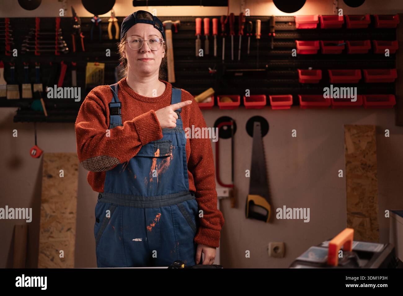 Guarda lì. La donna falegname indossa indumenti da lavoro in officina con attrezzi appesi al muro puntando il dito sullo spazio di copia . falegname donna in tarda serata Foto Stock