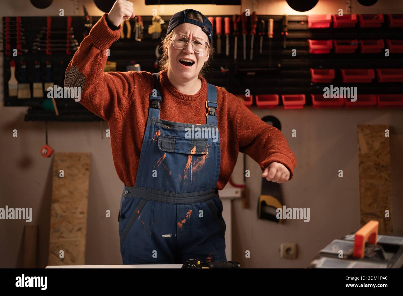 La donna falegname indossa indumenti da lavoro in officina con attrezzi appesi alla parete che scuote il pugno fissato alla telecamera minacciando. falegname donna in tarda serata Foto Stock