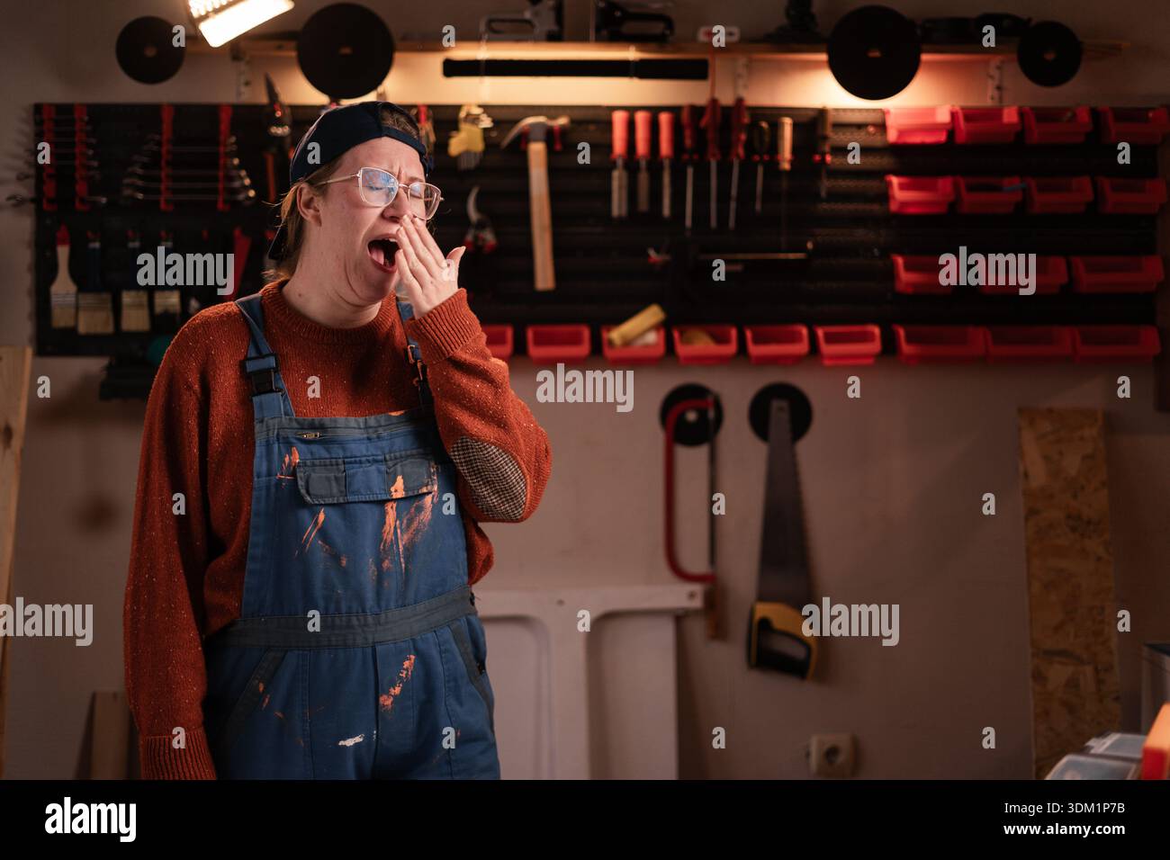 La donna falegname indossa indumenti da lavoro in officina con attrezzi appesi allo sbadiglio mentre posa davanti alla telecamera. falegname donna in tarda serata in autentica georgia Foto Stock