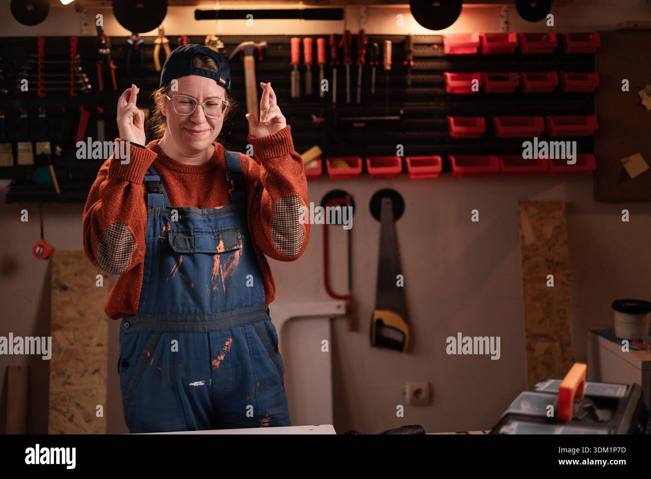 La donna falegname indossa abiti da lavoro in officina con gli attrezzi appesi al dito della parete. falegname donna in tarda serata in garage autentico. Foto Stock