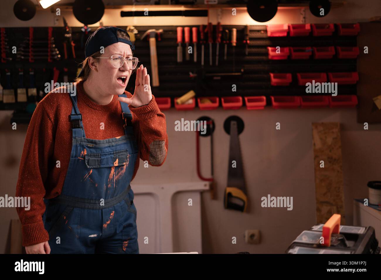 La donna falegname indossa indumenti da lavoro in officina con attrezzi appesi al muro che urla un annuncio. falegname donna in tarda serata in garage autentico. co Foto Stock