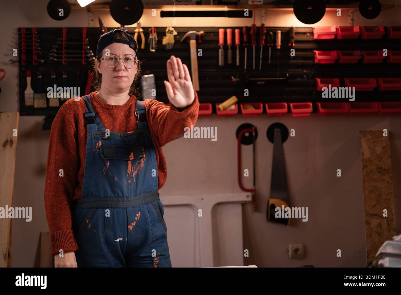 La donna falegname indossa indumenti da lavoro in officina con attrezzi appesi alla parete che fanno fermare il movimento guardando la fotocamera. falegname donna in tarda serata Foto Stock