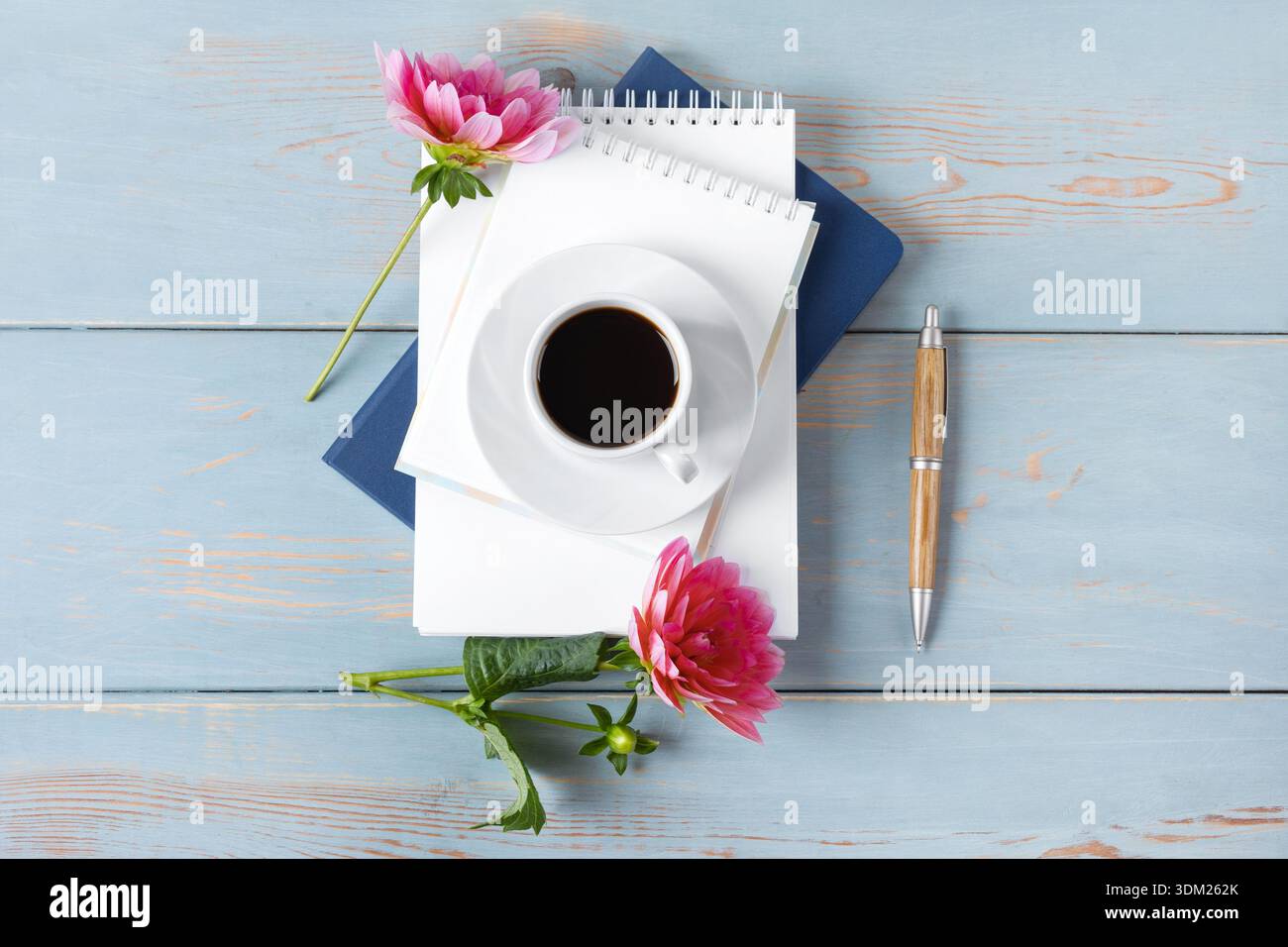 Vista dall'alto del desktop. Quaderno a spirale con pagina vuota aperta, penna, tazza bianca con caffè nero, fiori dahlia. Concetto di desktop per ufficio durante le festività. Tazza di caffè mattutina. Foto Stock