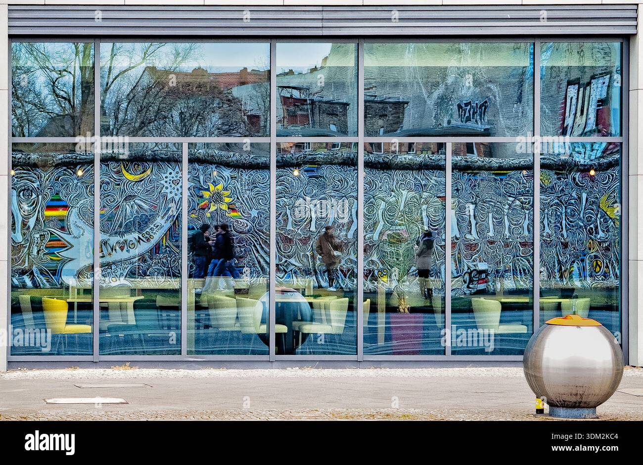 Riflessi dell'iconico muro dell'East Side Gallery Berlin Wall murals, con tonalità della bandiera tedesca, figure astratte, sole e luna, nelle grandi finestre di vetro di un caffè - Immagine stock catturata con smartphone