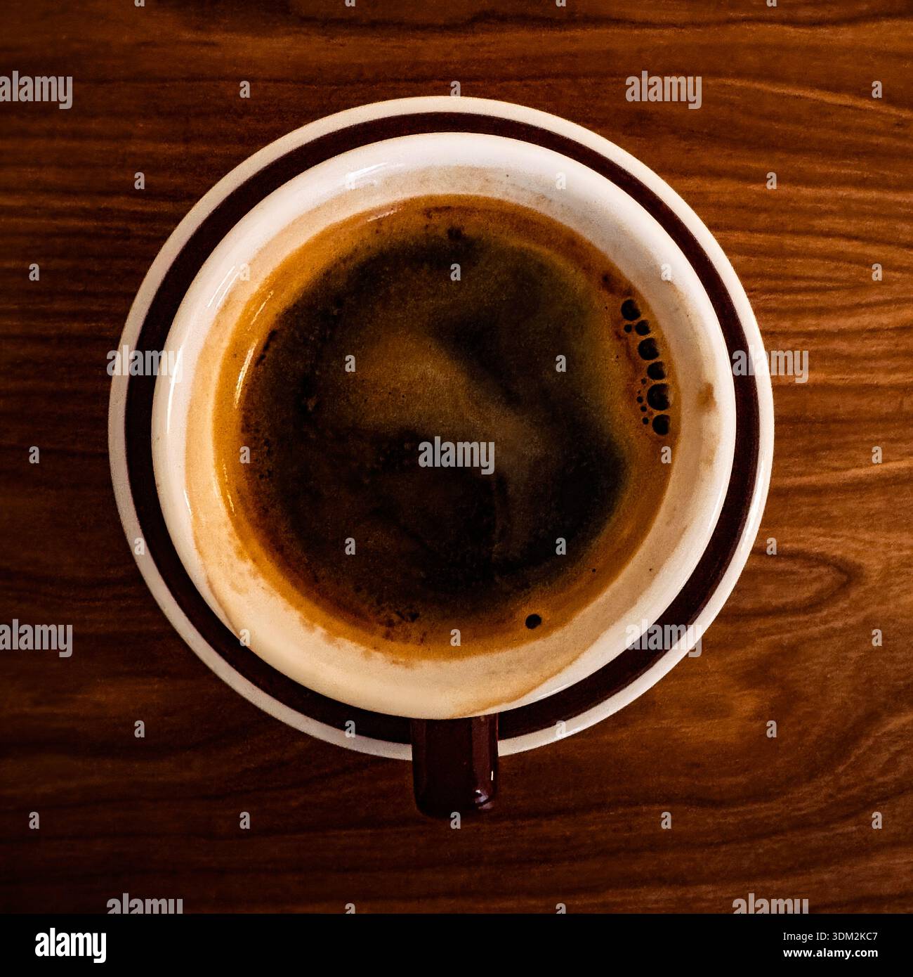 Vista dall'alto verso il basso di un espresso fresco in una tazza di porcellana bianca con spesse bolle di crema e schiuma su un piattino, posizionato su una calda superficie di legno - Immagine stock catturata con smartphone