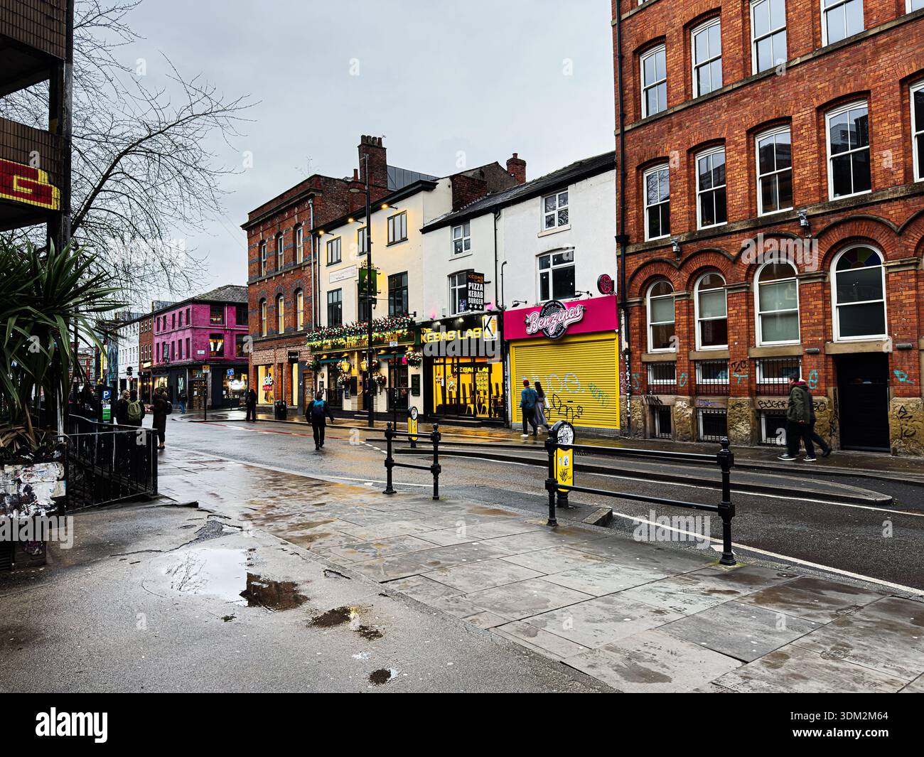 Manchester, Regno Unito - 21 gennaio 2026: Le vetrine colorate fiancheggiano una strada piovosa di Manchester con pedoni e vita urbana. Foto Stock