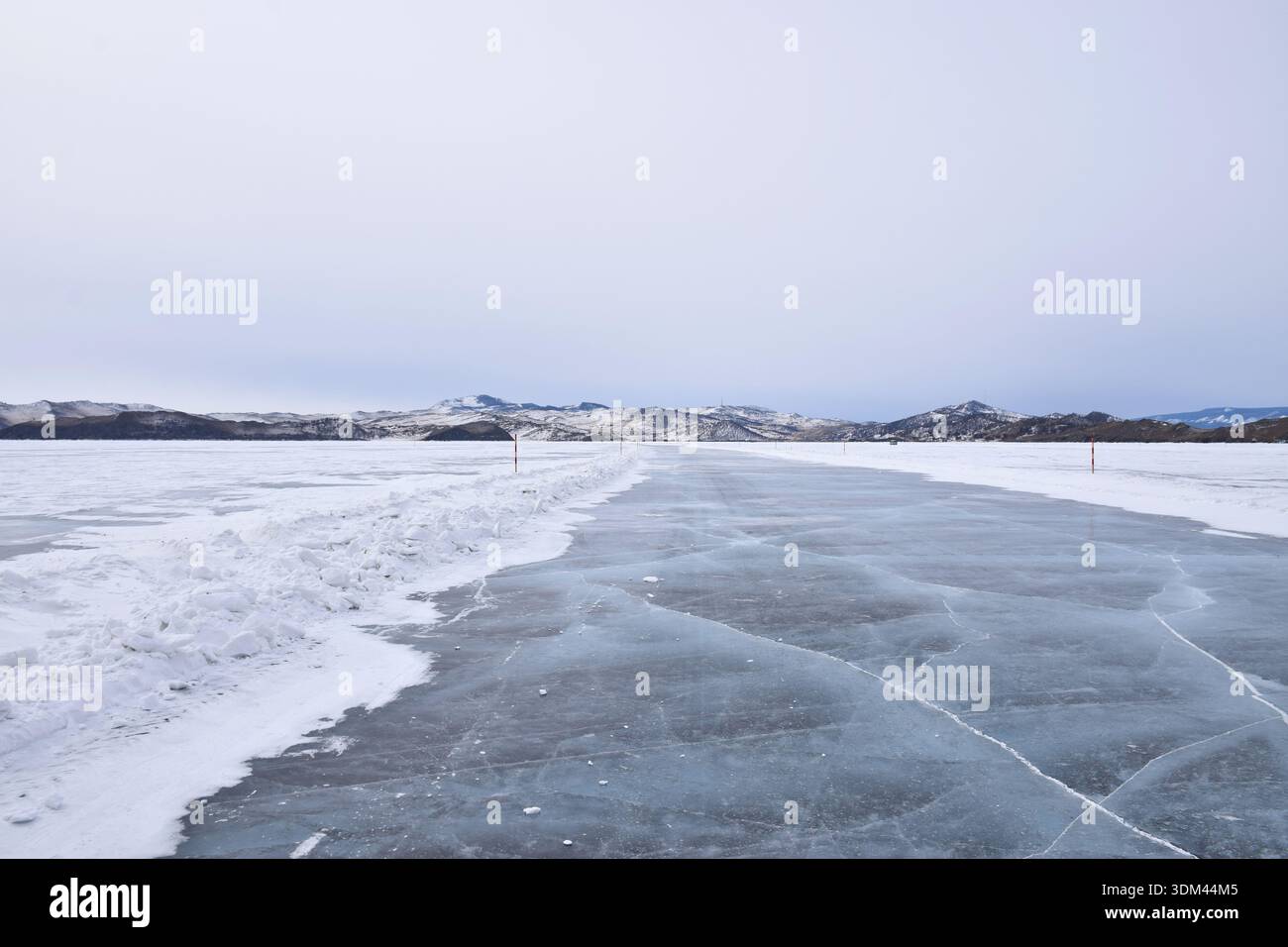 Strada invernale di ghiaccio per l'isola di Olkhon attraverso il lago ghiacciato Baikal, Siberia. Superficie ghiacciata ricoperta di neve con crepe, lontane montagne innevate sotto il cielo coperto. Foto Stock