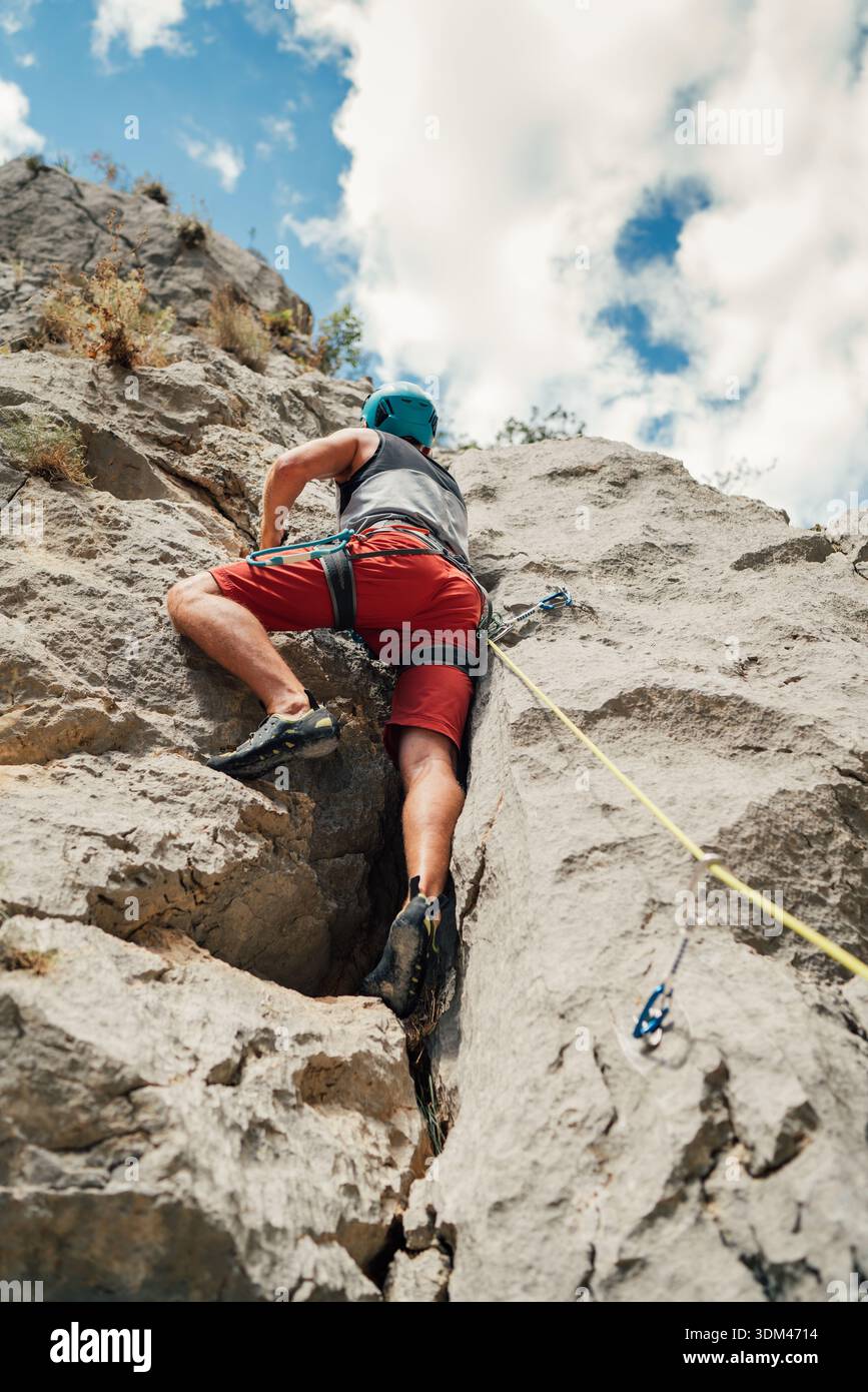 Arrampicatore maschile in pantaloncini rossi e casco blu che arrampicano sulla ripida parete di montagna a Paklenica, Croazia. Atleta professionista che utilizza corda e paraspruzzi rapidi Foto Stock