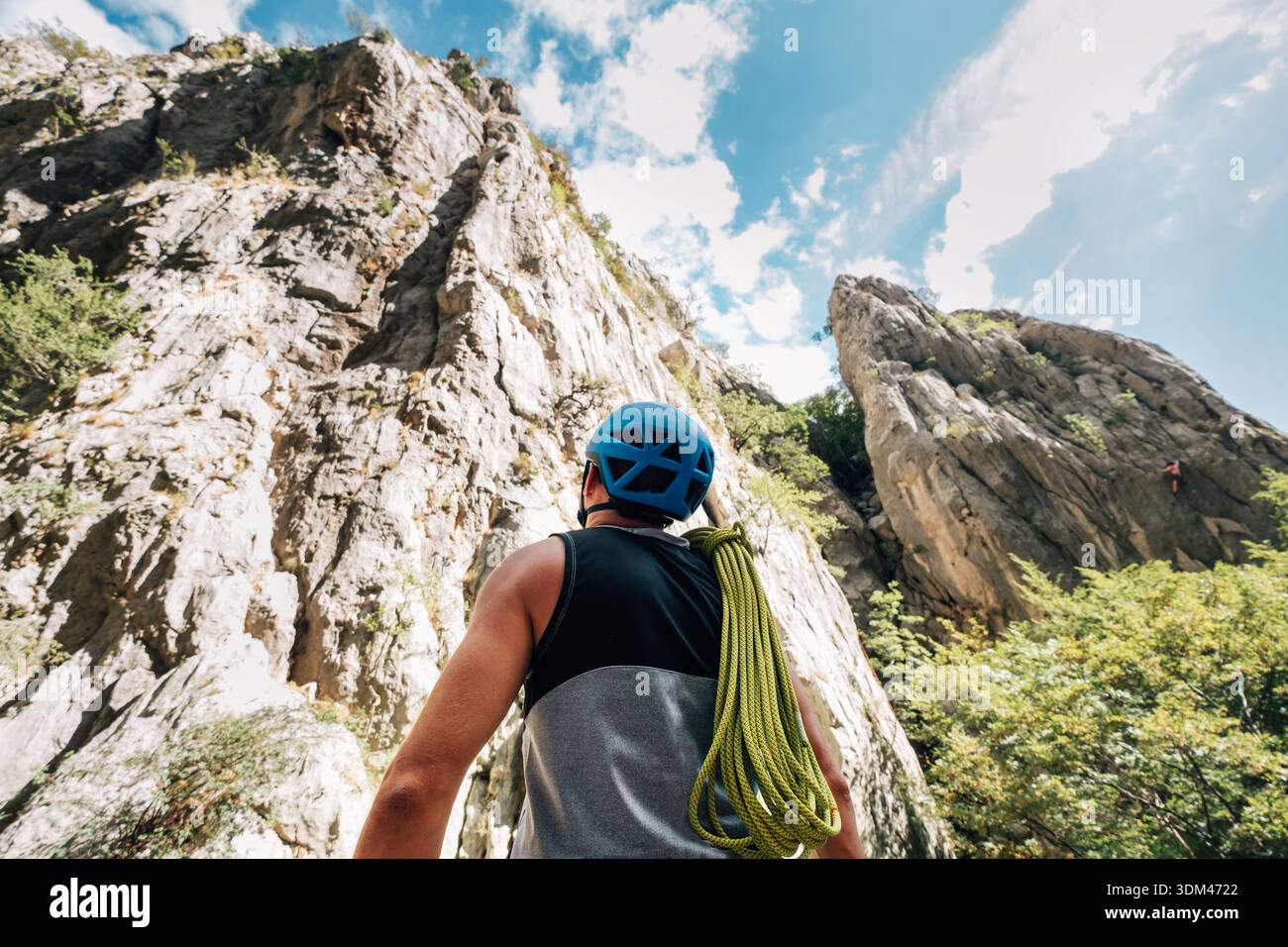 Vista posteriore di un alpinista maschio con casco blu e corda verde che guarda l'enorme parete della scogliera. Alpinista professionista che si prepara per gli sport estremi Foto Stock