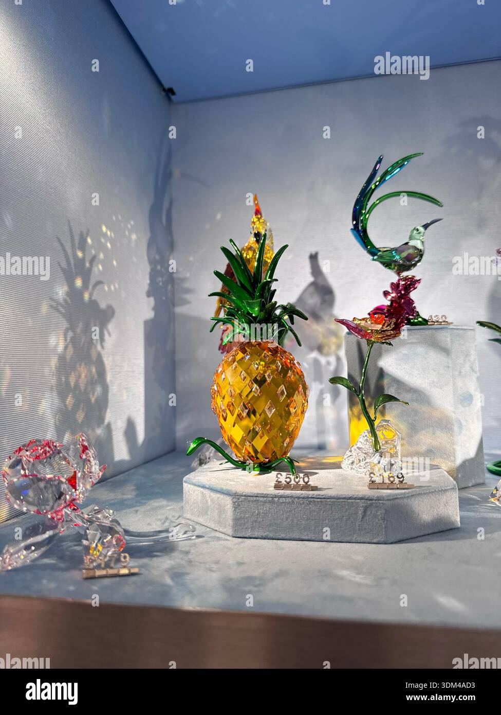 Londra, Regno Unito, 5 dicembre 2025, collezione di statuette in cristallo con ananas giallo oro con foglie verdi, uccello colorato, fiori su bianco Foto Stock